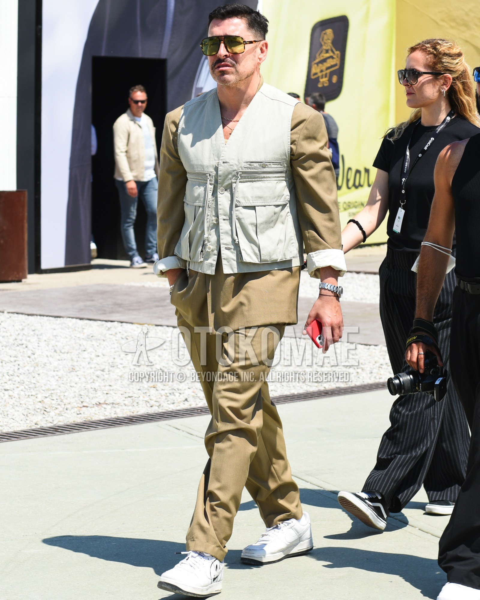 Men's spring summer outfit with brown tortoiseshell sunglasses, olive green plain casual vest, white low-cut sneakers, beige plain suit.