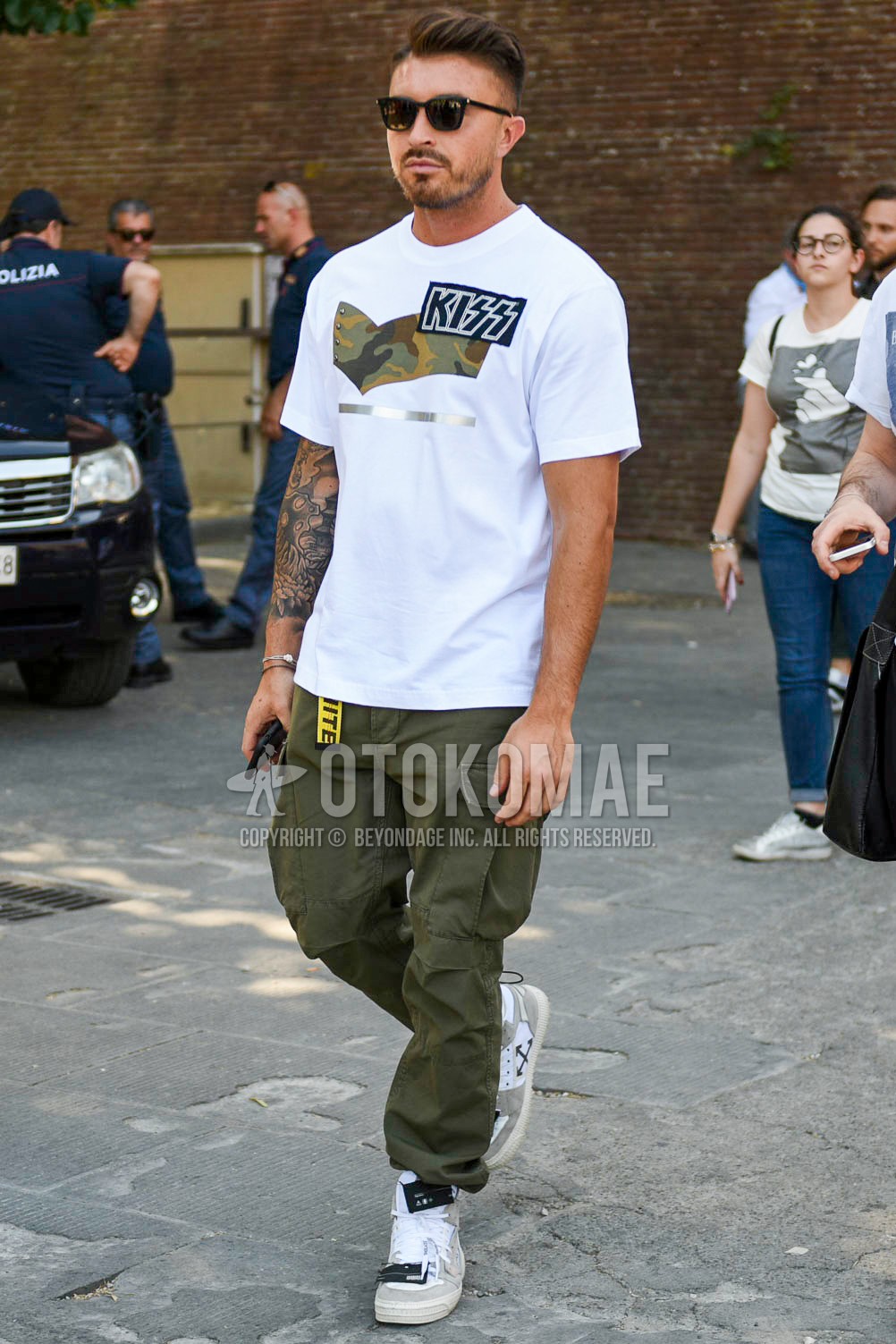 Men's summer outfit with black plain sunglasses, white graphic t-shirt, yellow graphic tape belt, olive green plain cargo pants, gray high-cut sneakers.