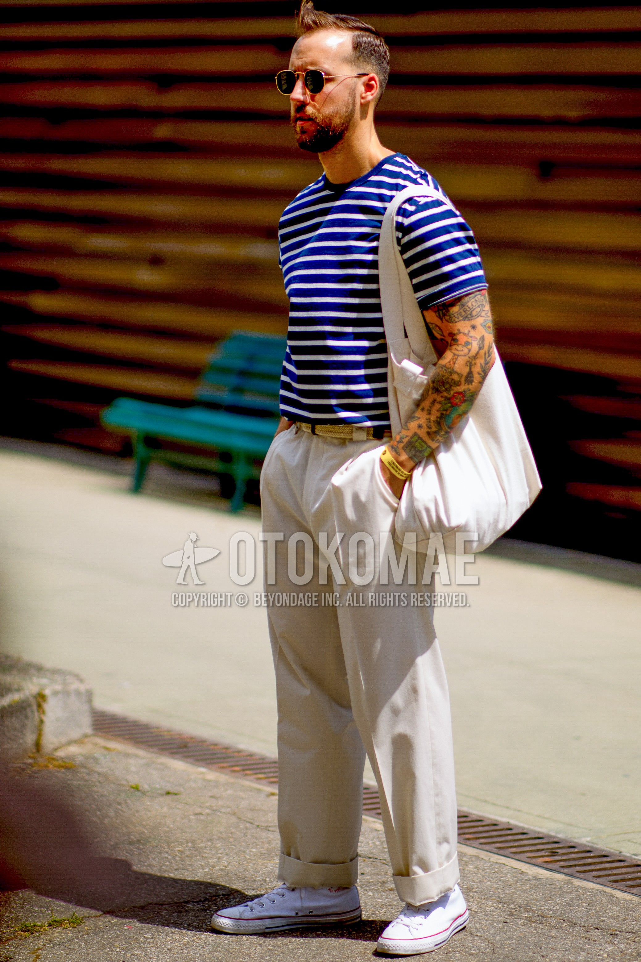 Men's spring summer outfit with gold plain sunglasses, blue horizontal stripes t-shirt, white plain braided belt, white plain slacks, white low-cut sneakers, white plain tote bag.