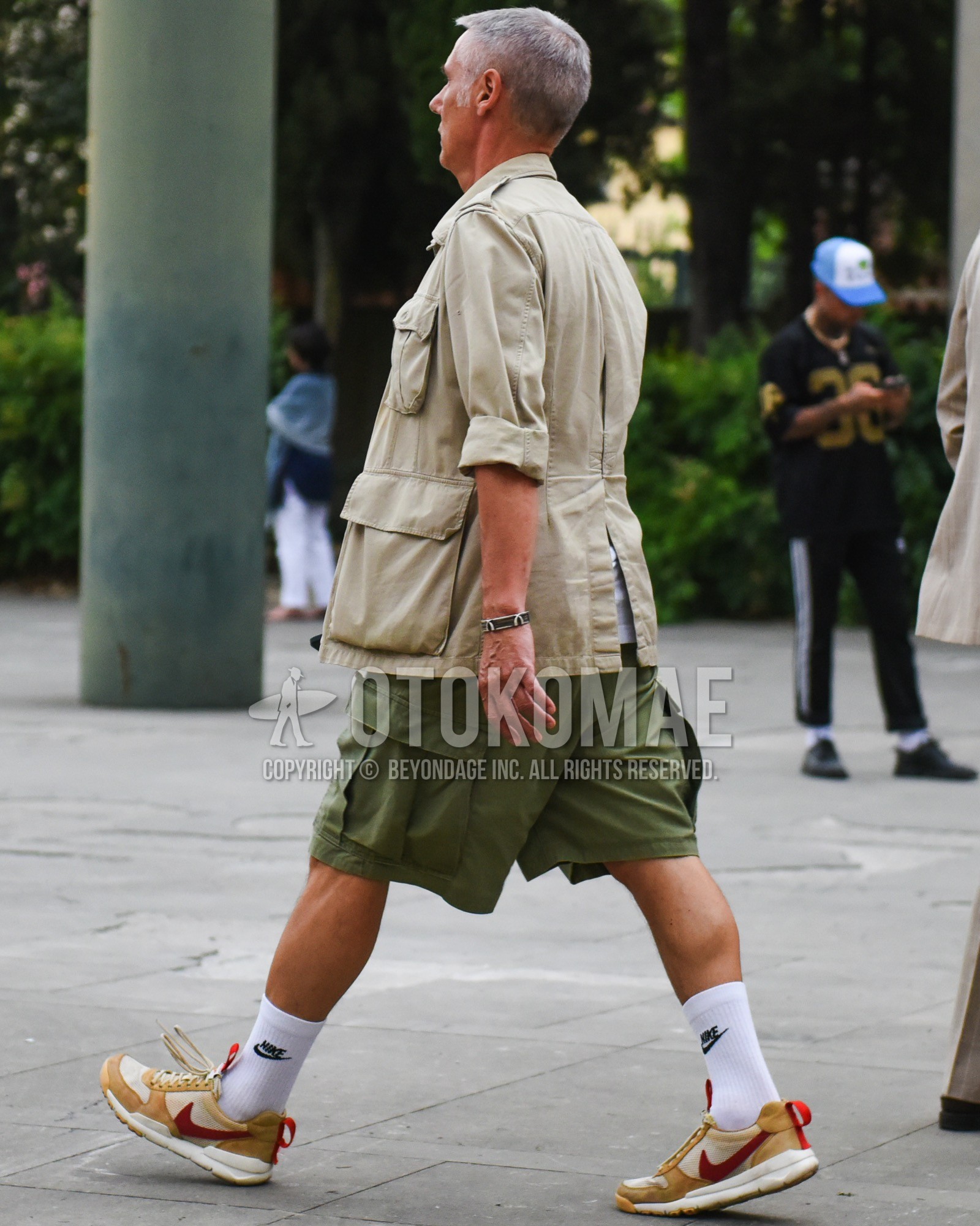 Men's spring summer outfit with beige plain safari jacket, olive green plain short pants, olive green plain cargo pants, white one point socks, olive green low-cut sneakers.