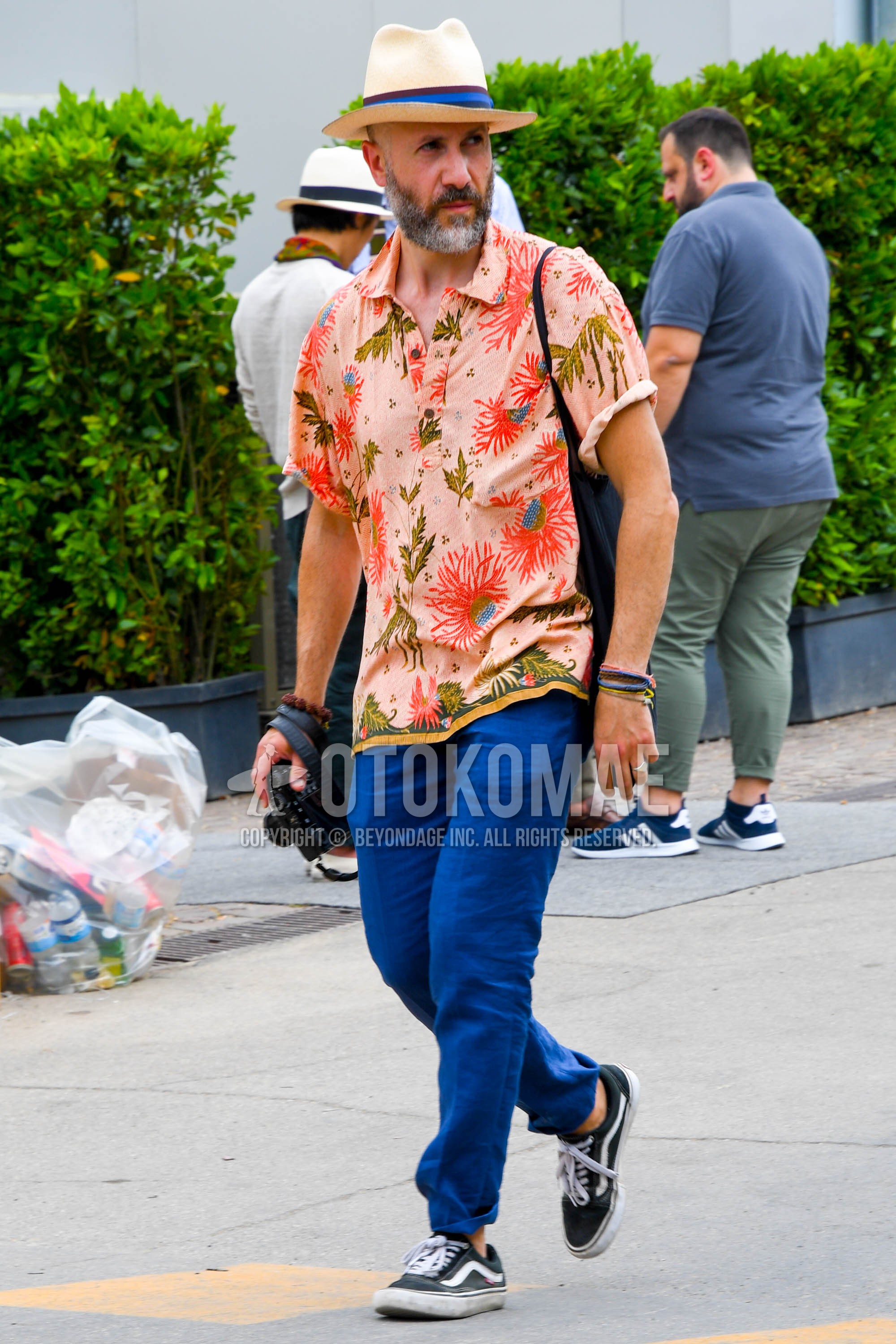 Men's summer outfit with beige plain hat, orange botanical shirt, black low-cut sneakers.