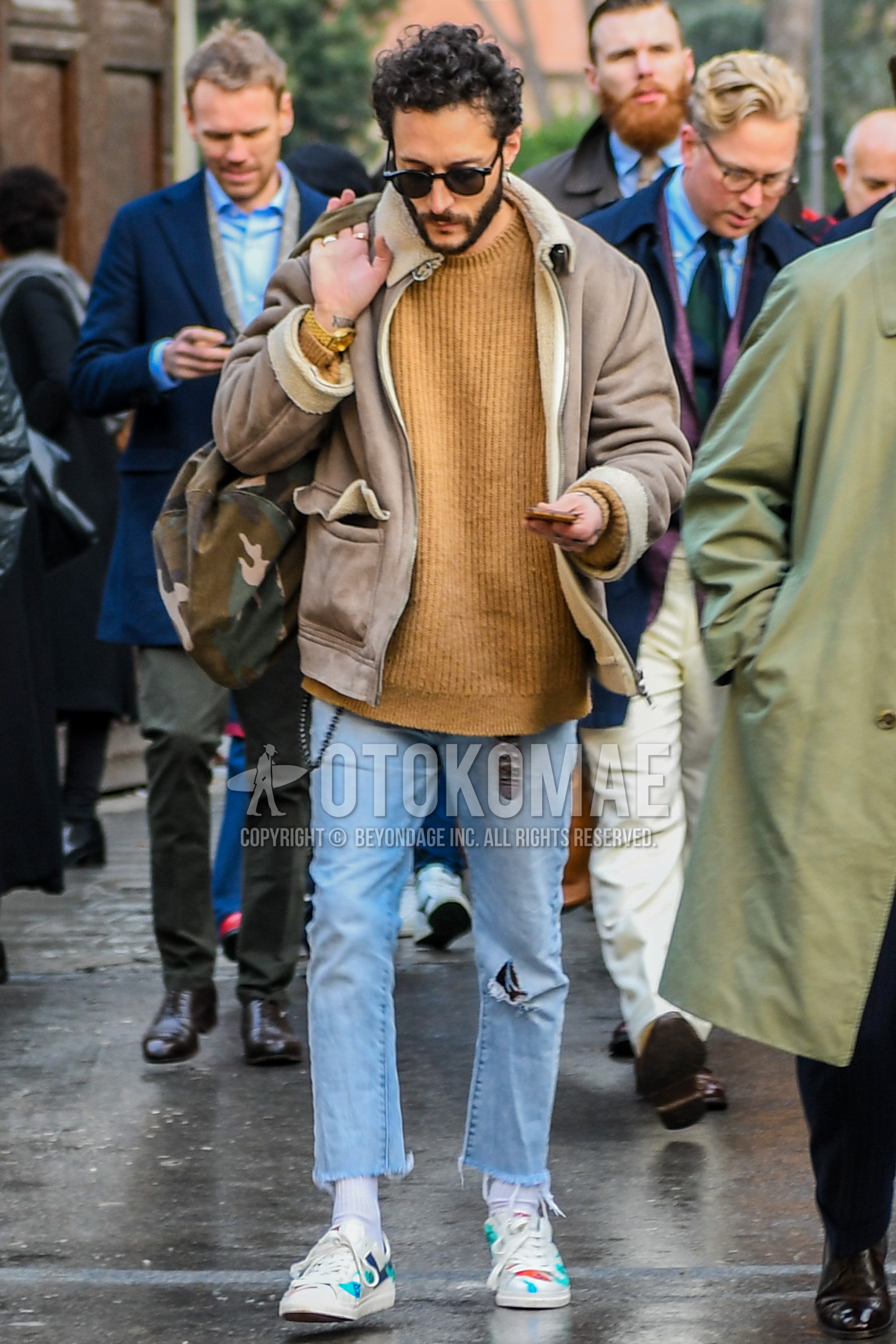 Men's winter outfit with plain sunglasses, brown plain leather jacket, brown plain sweater, light blue plain damaged jeans, white plain socks, multi-color low-cut sneakers.