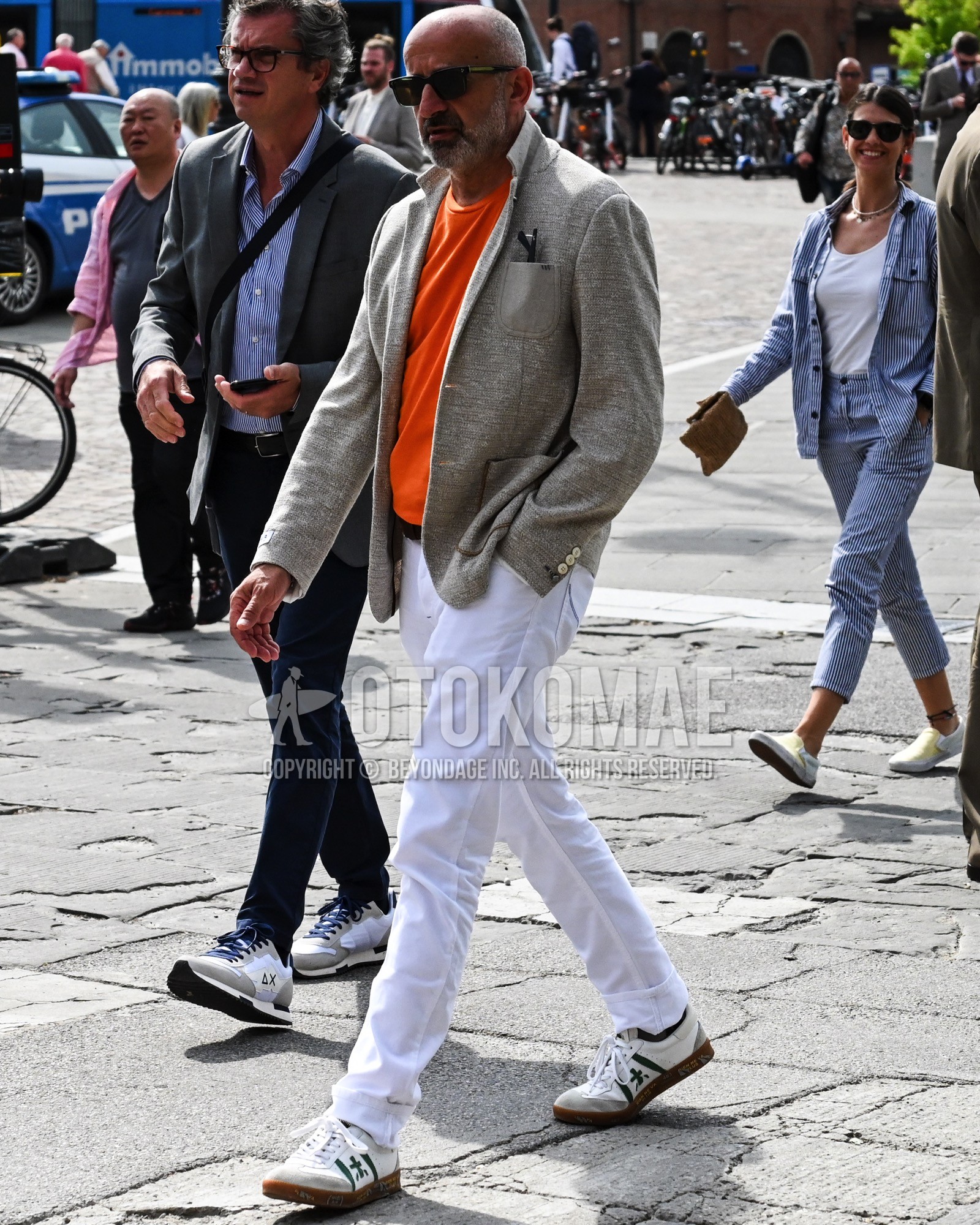 Men's spring summer autumn outfit with black plain sunglasses, gray plain tailored jacket, orange plain t-shirt, white plain chinos, white low-cut sneakers.