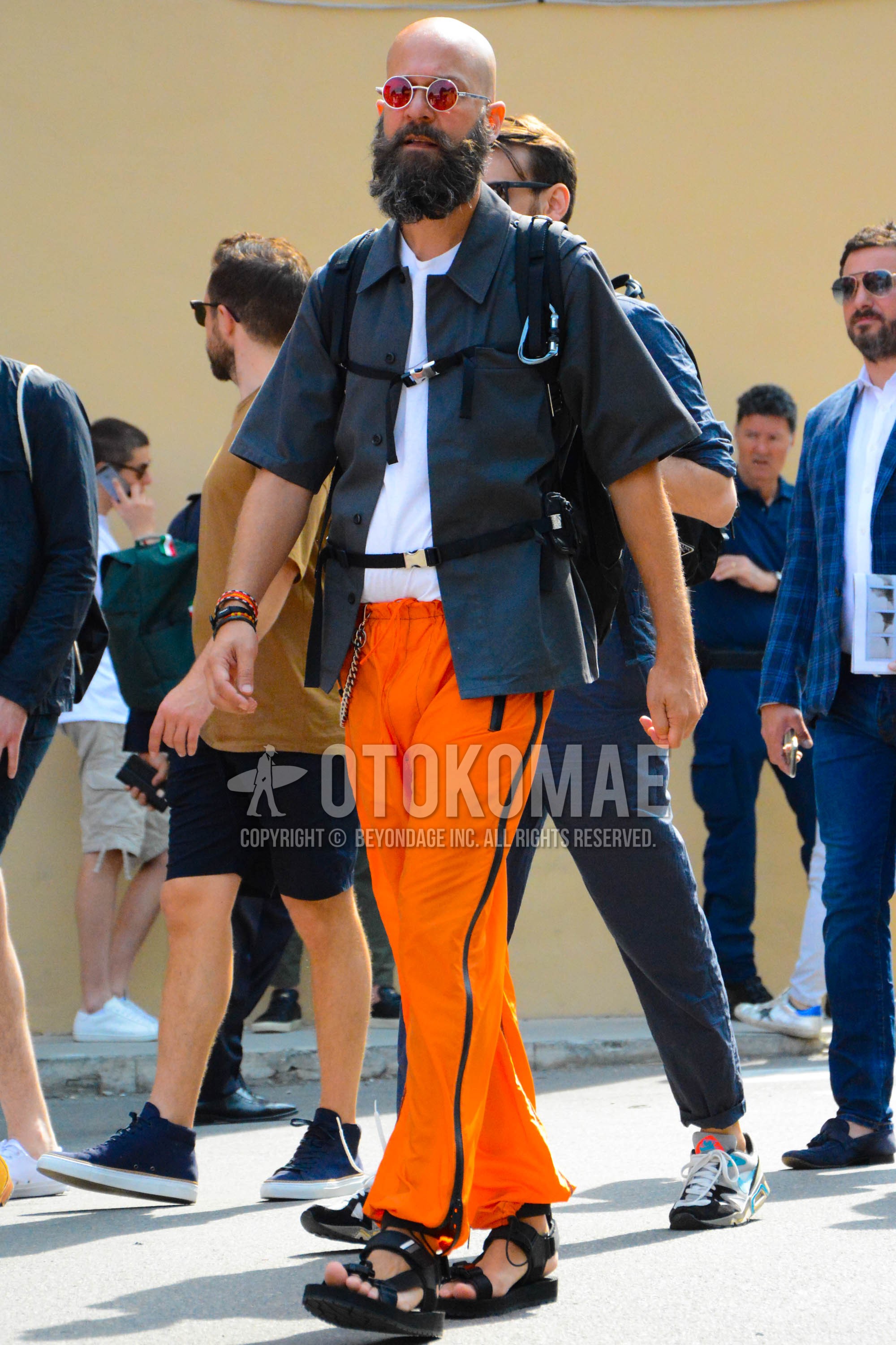 Men's summer outfit with silver plain sunglasses, white plain t-shirt, gray plain shirt, orange plain sideline pants, black sport sandals, black plain backpack.