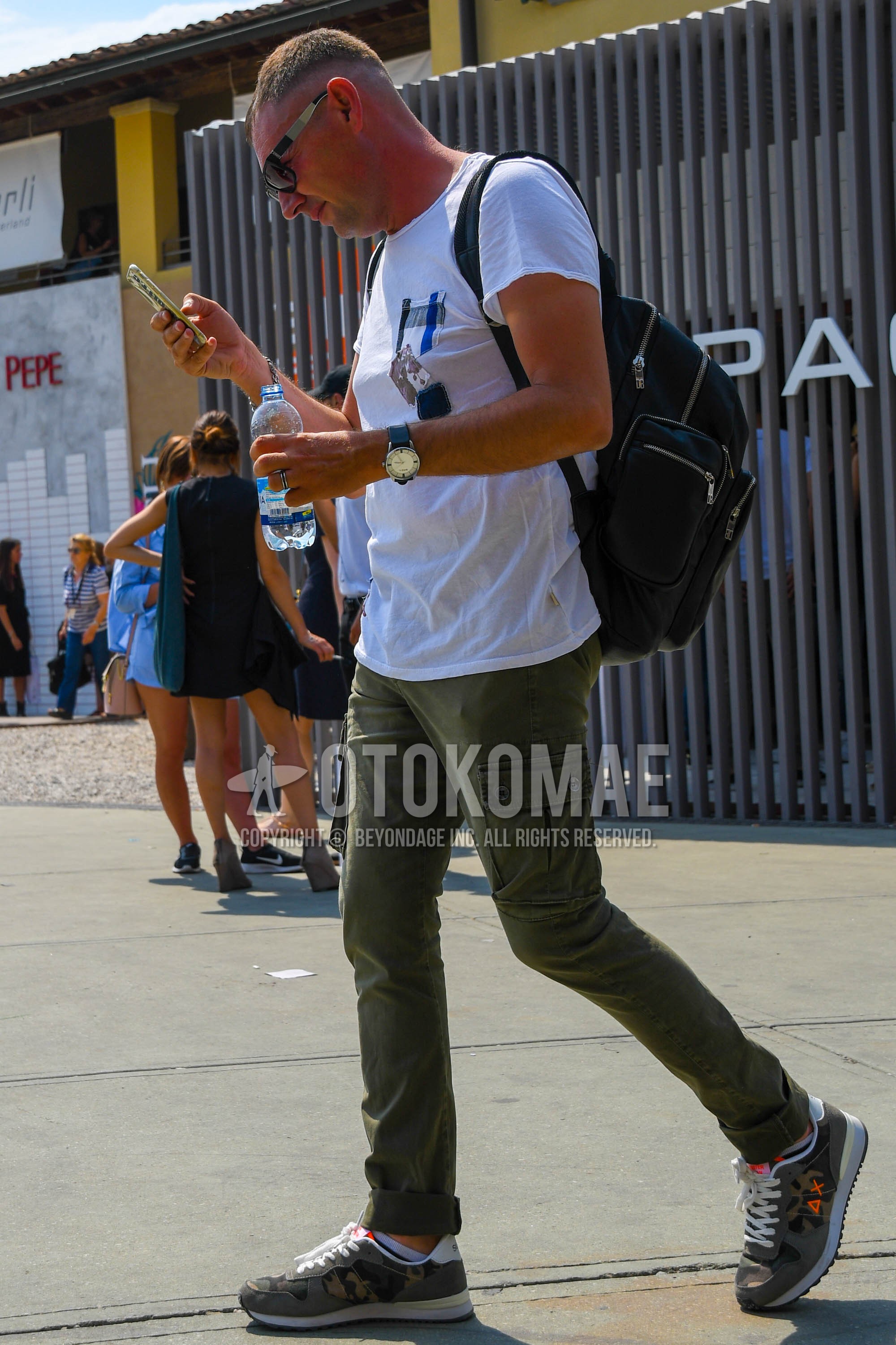 Men's spring summer outfit with plain sunglasses, white graphic t-shirt, olive green plain cargo pants, olive green low-cut sneakers, black plain backpack.