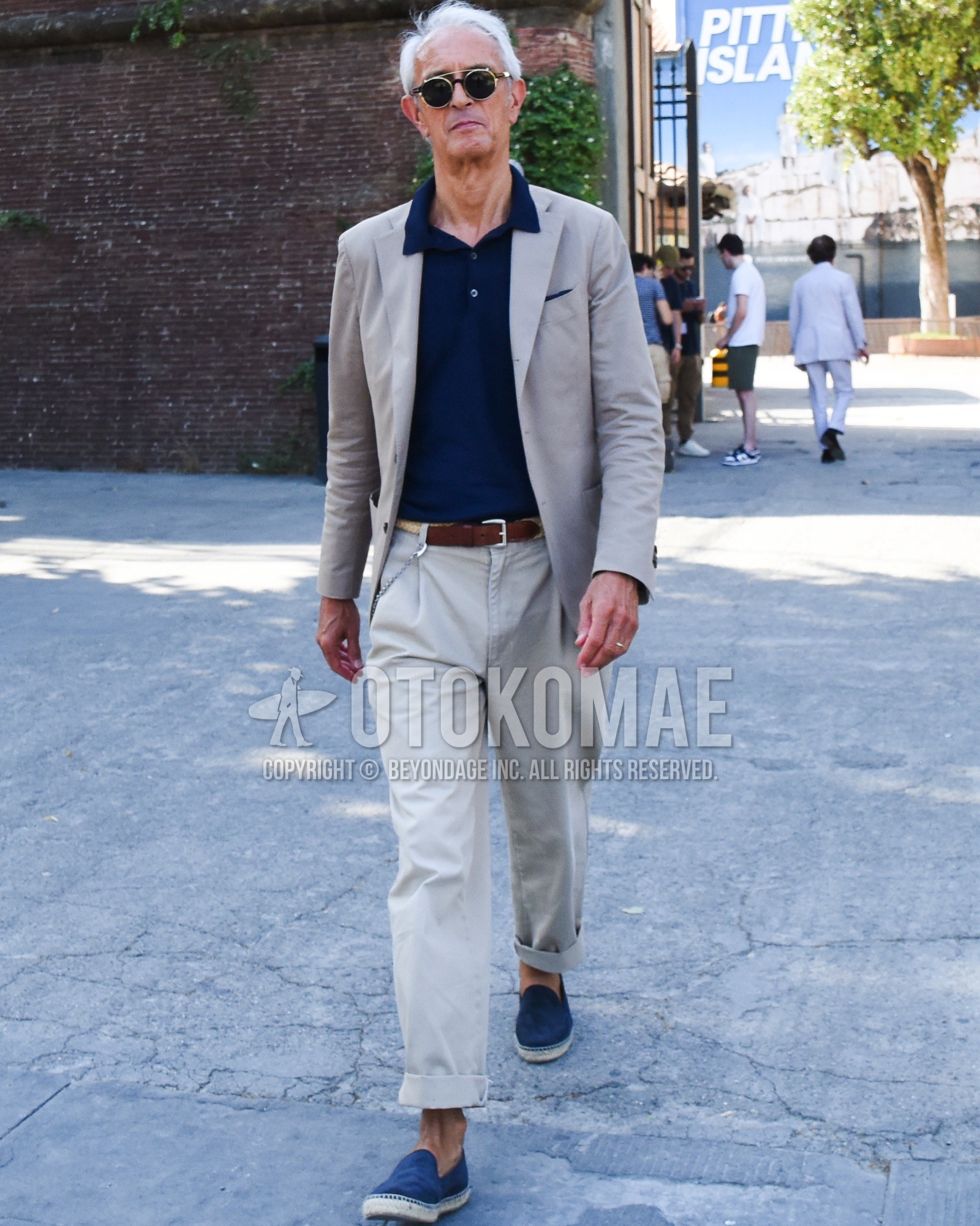 Men's spring summer outfit with gold plain sunglasses, navy plain polo shirt, brown plain leather belt, navy slip-on sneakers, gray plain suit.