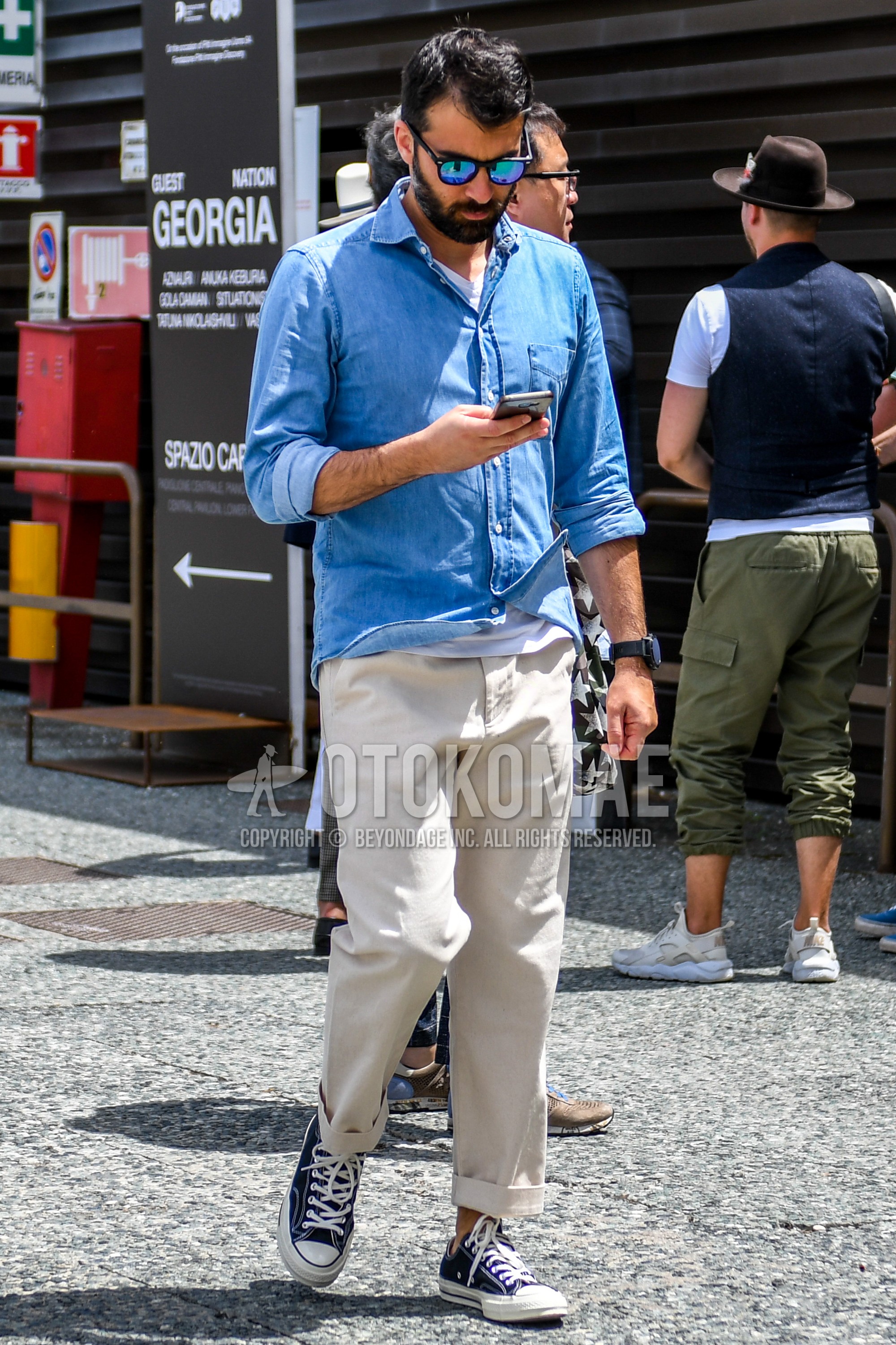 Men's spring summer autumn outfit with plain sunglasses, blue plain shirt, white plain t-shirt, beige plain cotton pants, navy low-cut sneakers.