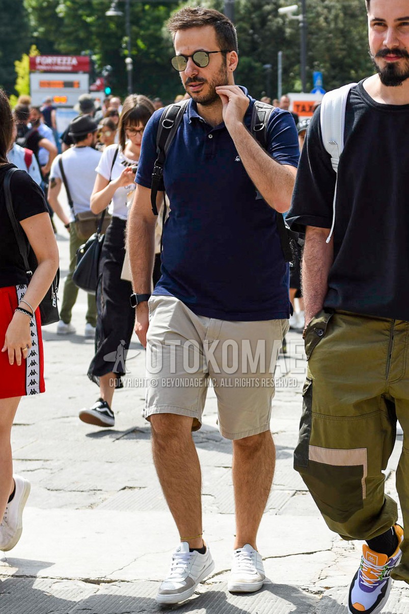 Men's summer outfit with black plain sunglasses, navy plain polo shirt, beige plain chinos, plain short pants, white low-cut sneakers.