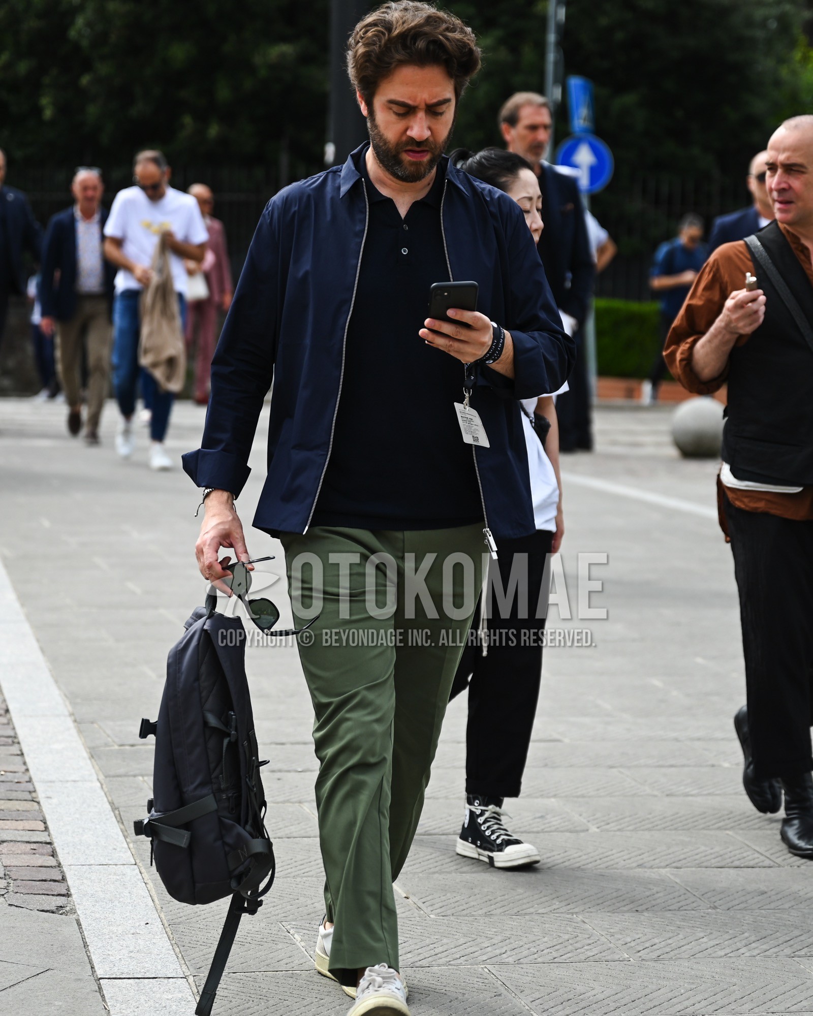 Men's spring summer autumn outfit with navy plain swing top, black plain t-shirt, olive green plain chinos, white low-cut sneakers, black plain backpack.