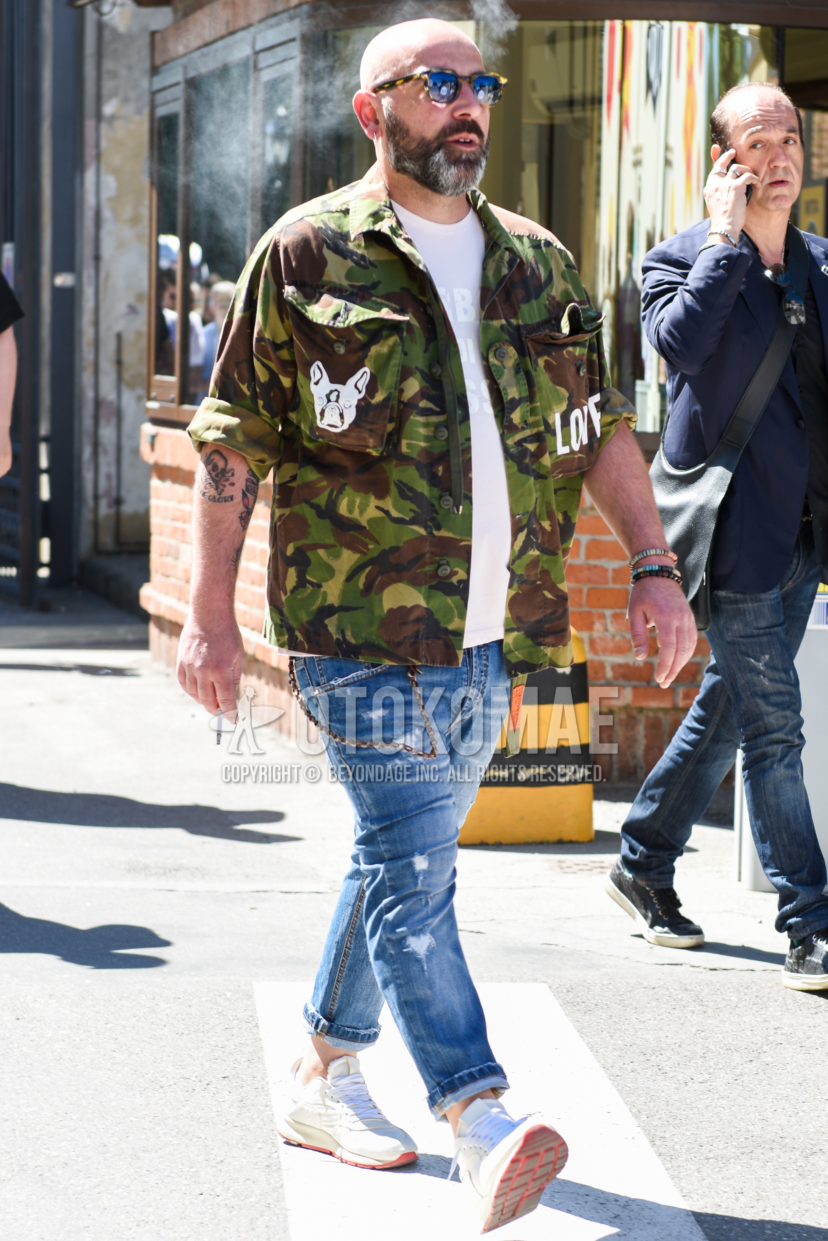 Men's spring autumn outfit with beige tortoiseshell sunglasses, olive green brown beige camouflage shirt jacket, white graphic t-shirt, blue plain damaged jeans, beige low-cut sneakers.