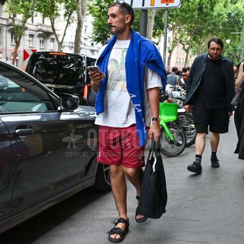 黒無地のサングラス、白グラフィックのTシャツ、ブルーレタードのロンT、レッドボトムスのショートパンツ、黒スポーツサンダル、黒無地のブリーフケース/ハンドバッグを合わせた夏のメンズコーデ・着こなし。