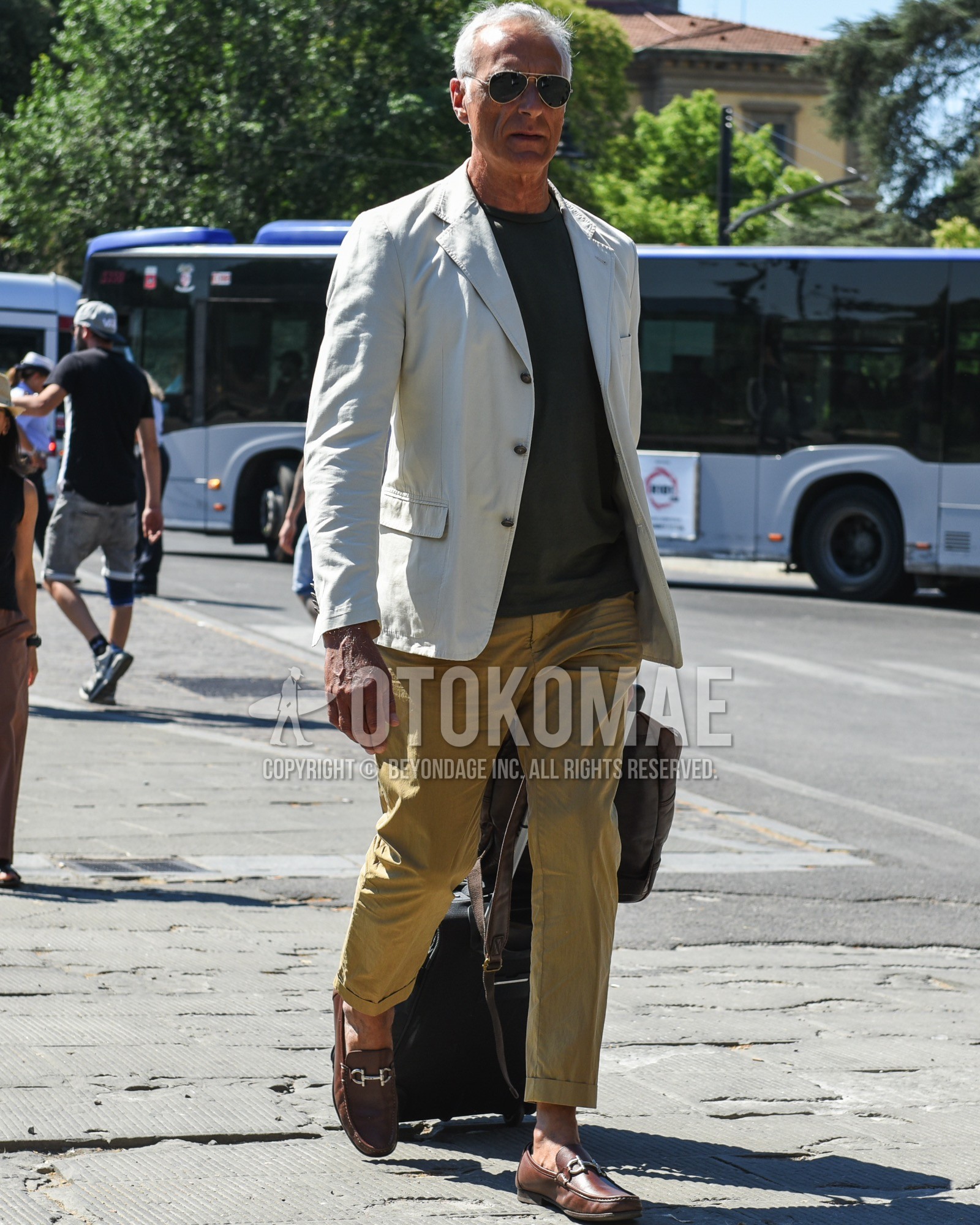 Men's spring summer outfit with gold plain sunglasses, white plain tailored jacket, olive green plain t-shirt, beige plain chinos, brown bit loafers leather shoes, black plain suitcase.