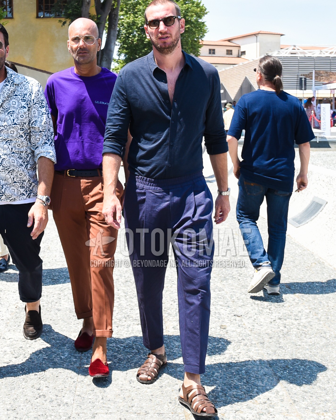 Men's spring summer outfit with black plain sunglasses, navy plain shirt, purple plain easy pants, purple plain pleated pants, brown gurkha sandals.