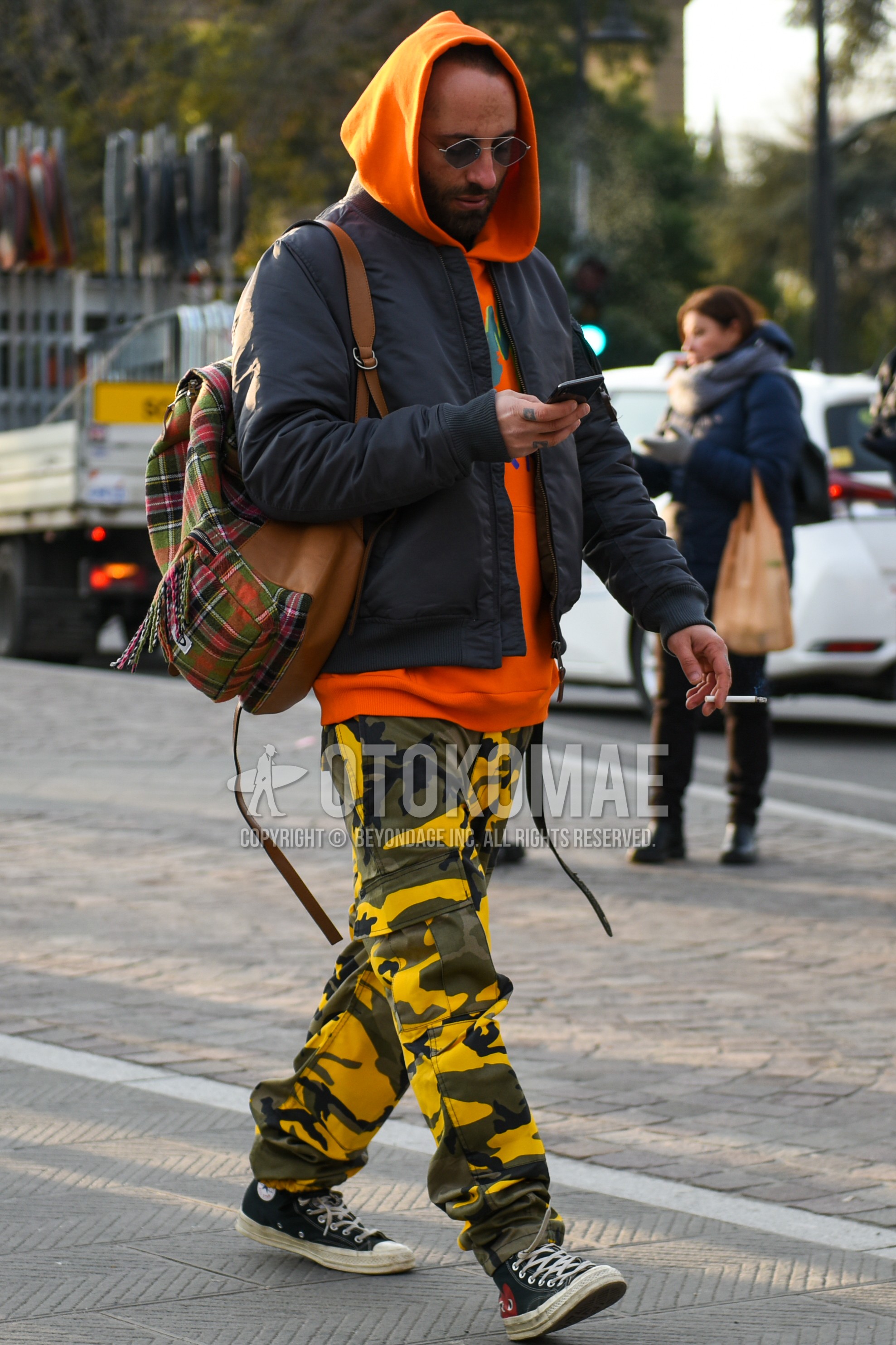 Men's autumn winter outfit with silver plain sunglasses, gray plain MA-1, orange graphic hoodie, yellow gray camouflage cargo pants, black high-cut sneakers, multi-color brown check backpack.