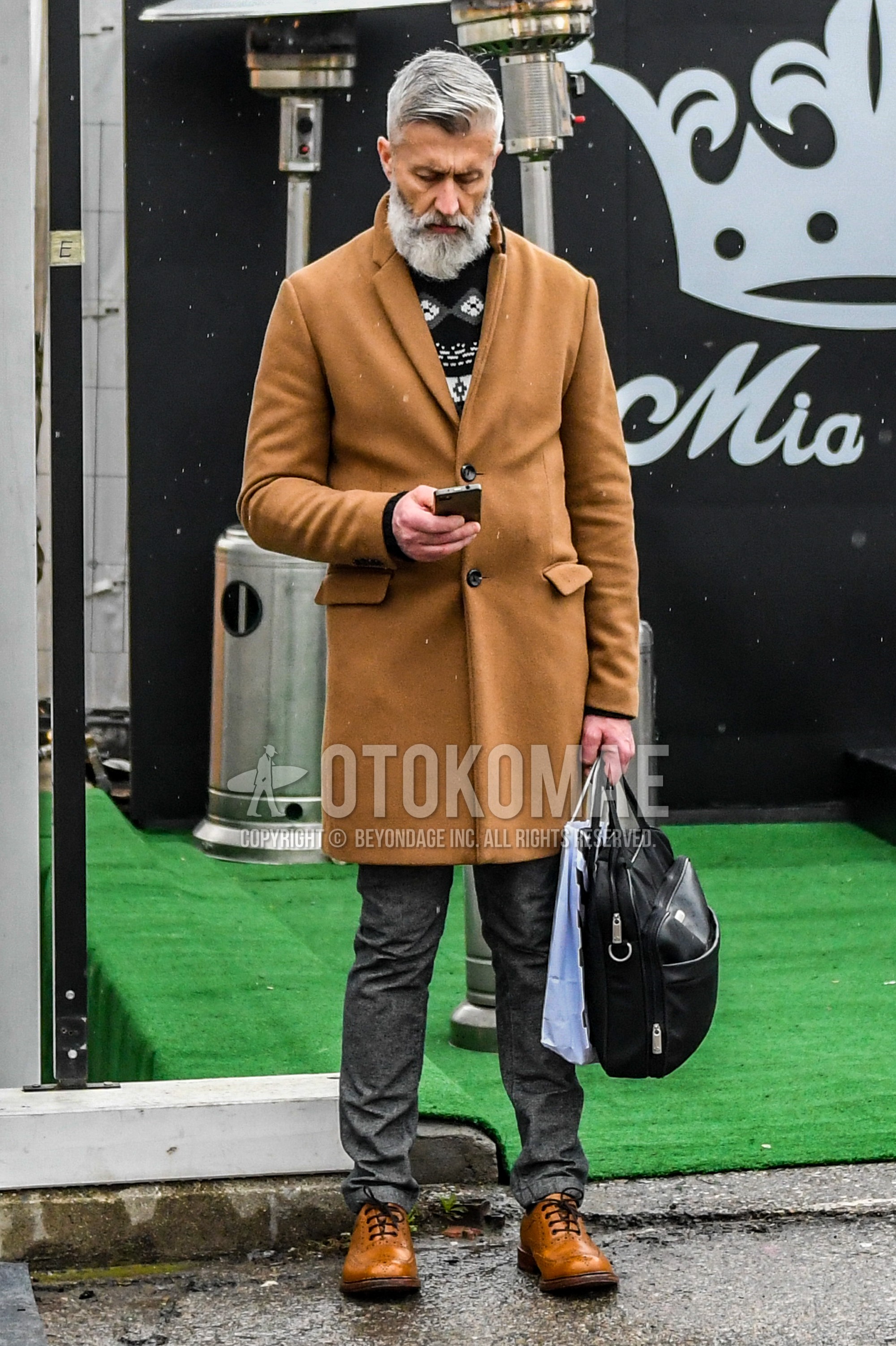 Men's autumn winter outfit with brown plain chester coat, black graphic sweater, gray plain slacks, beige wing-tip shoes leather shoes, black plain briefcase/handbag.