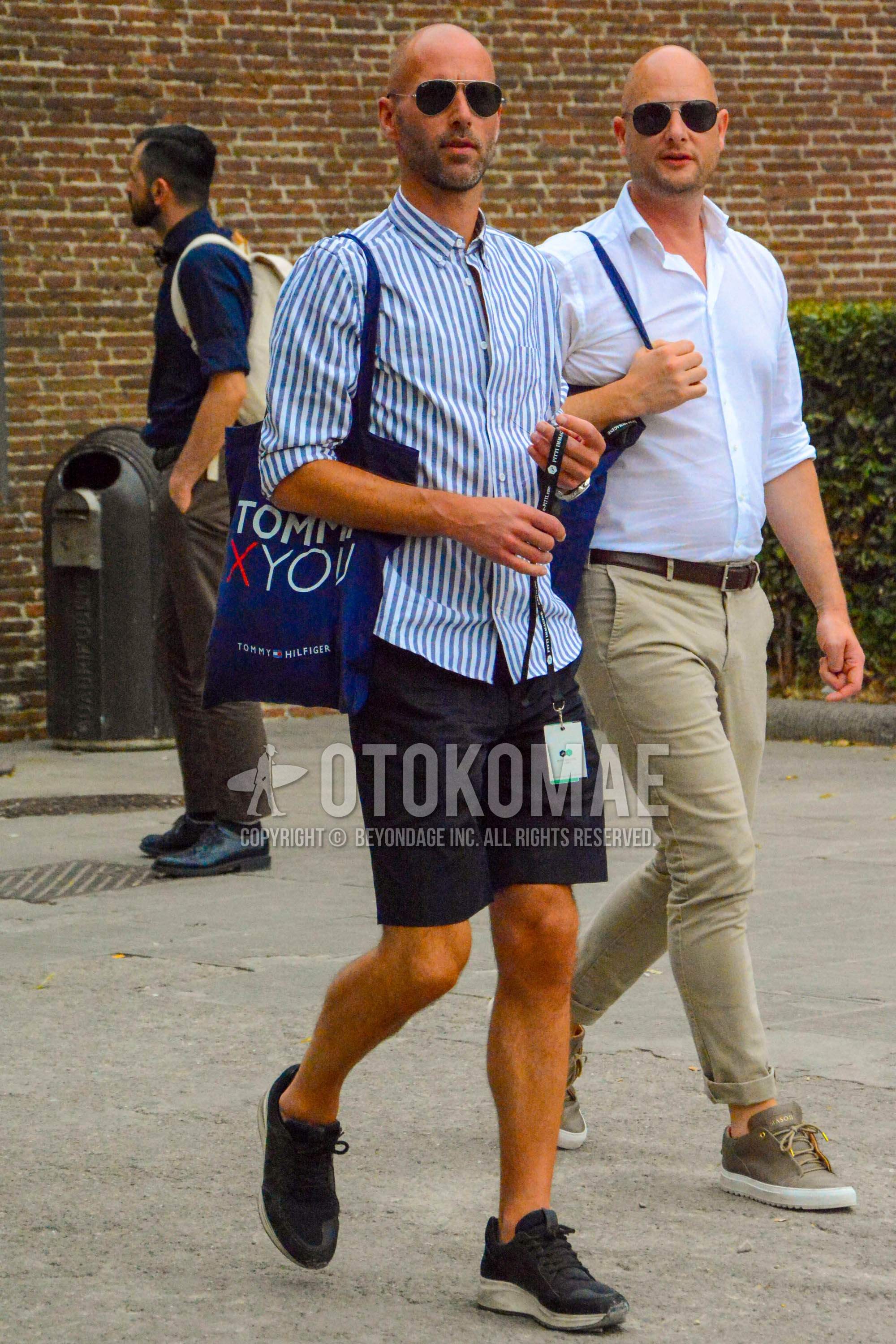 Men's summer outfit with plain sunglasses, light blue white stripes shirt, navy plain short pants, black low-cut sneakers, blue graphic tote bag.