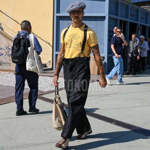 グレー無地のハンチングキャップ / キャスケット、ボストンの黒無地のメガネ、イエローレタードのTシャツ、黒無地のサスペンダー、黒無地のデニム/ジーンズ、ブラウン・白のブーツ、ベージュ無地のバッグを合わせた夏春のメンズコーデ・着こなし。