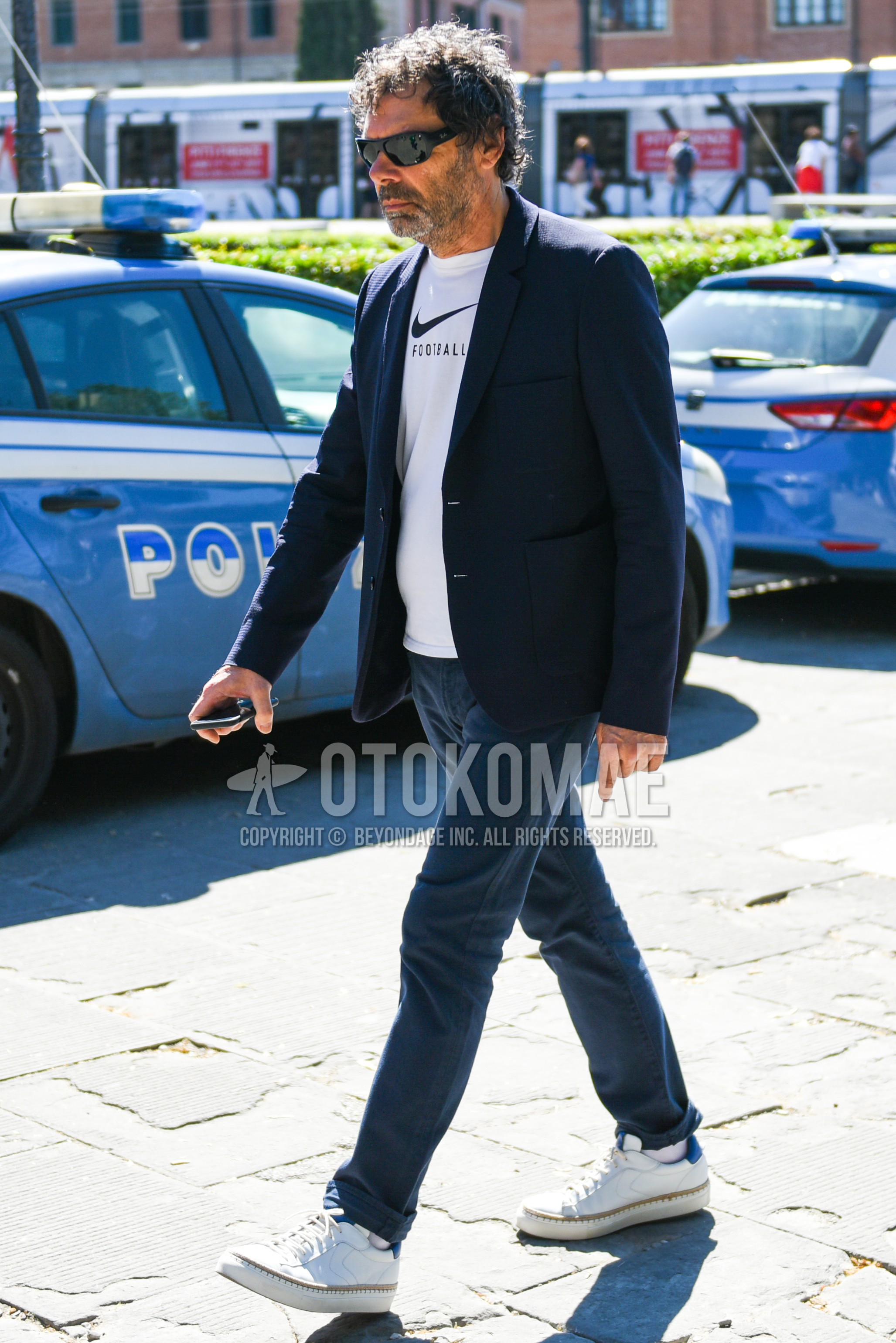 Men's spring summer autumn outfit with black plain sunglasses, navy plain tailored jacket, white graphic t-shirt, navy plain chinos, white low-cut sneakers.