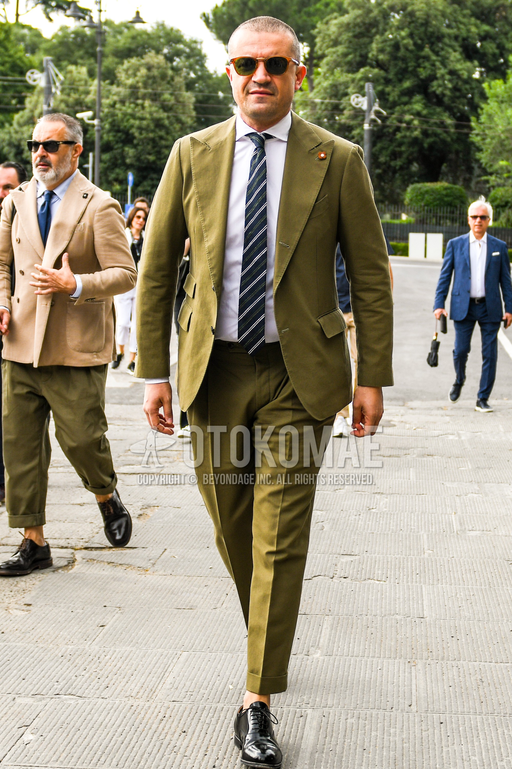 Men's spring autumn outfit with brown tortoiseshell sunglasses, white plain shirt, black straight-tip shoes leather shoes, olive green plain suit, navy green stripes necktie.