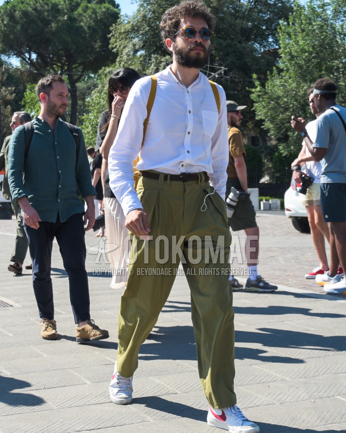Men's spring summer outfit with brown tortoiseshell sunglasses, white plain shirt, brown plain leather belt, olive green plain cotton pants, olive green plain pleated pants, white low-cut sneakers, yellow plain backpack.