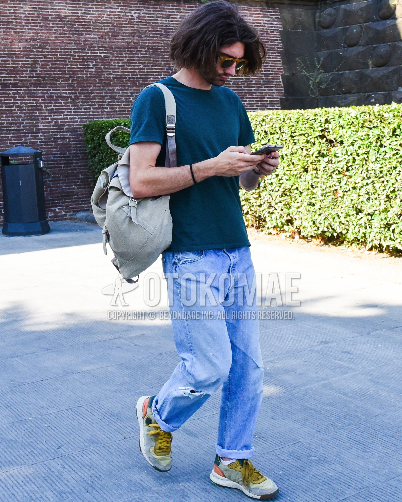Men's spring summer outfit with yellow plain sunglasses, green plain t-shirt, blue plain damaged jeans, gray low-cut sneakers, gray plain backpack.