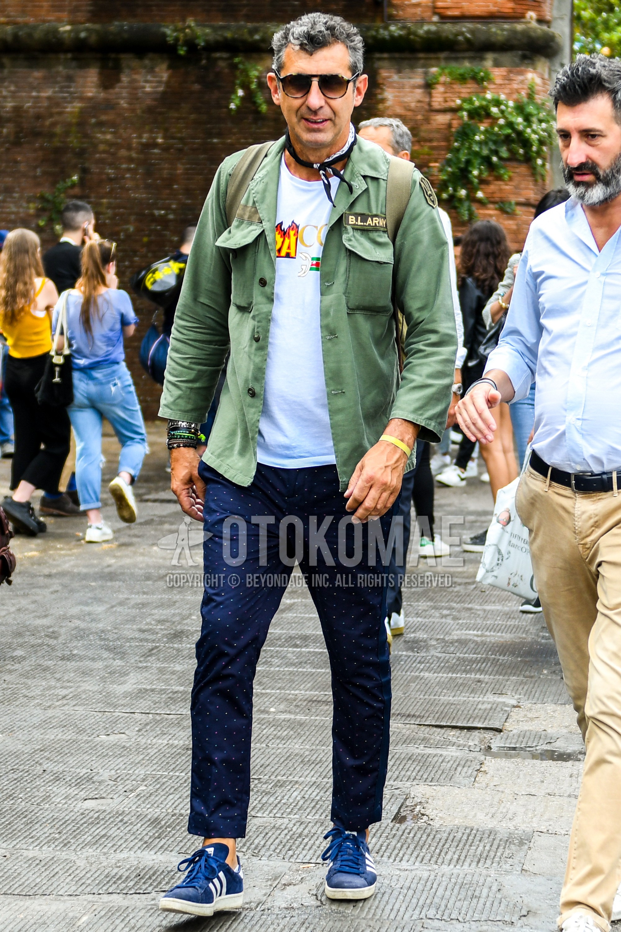 Men's spring summer autumn outfit with beige tortoiseshell sunglasses, black paisley bandana/neckerchief, olive green plain shirt jacket, white graphic t-shirt, navy dots chinos, blue low-cut sneakers.