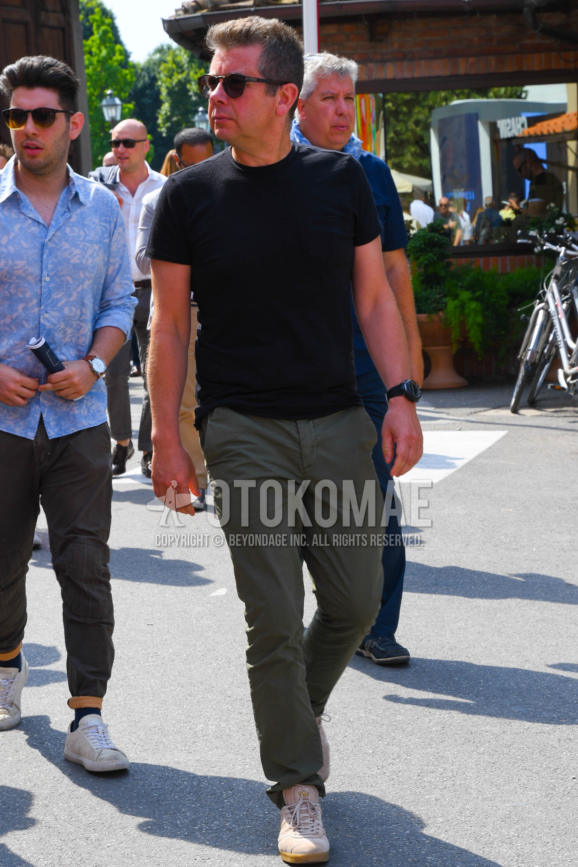 Men's spring summer outfit with black plain sunglasses, black plain t-shirt, olive green plain chinos, beige low-cut sneakers.