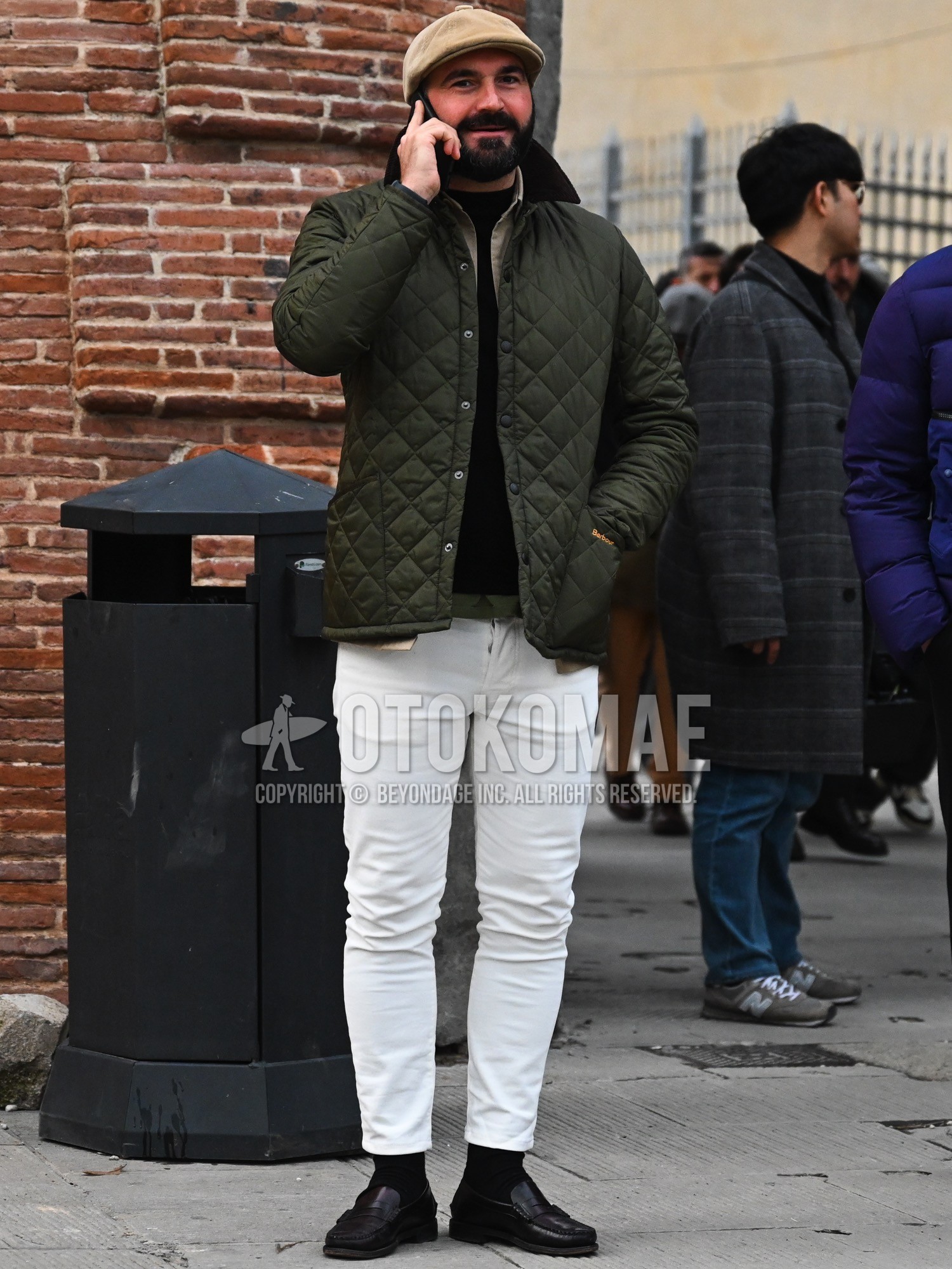 Men's autumn winter outfit with beige plain hunting cap, olive green plain quilted jacket, beige plain shirt, olive green plain t-shirt, black plain sweater, white plain cotton pants, white plain ankle pants, black plain socks, brown coin loafers leather shoes.