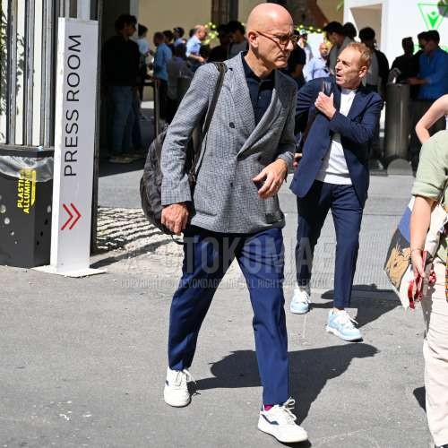 オレンジ無地のメガネ、グレーチェックのテーラードジャケット、ネイビー無地のポロシャツ、ブルー無地のスラックス、レッド無地のソックス、白・ブルーローカットのスニーカー、グレー・黒無地のバックパックを合わせた春秋のメンズコーデ・着こなし。