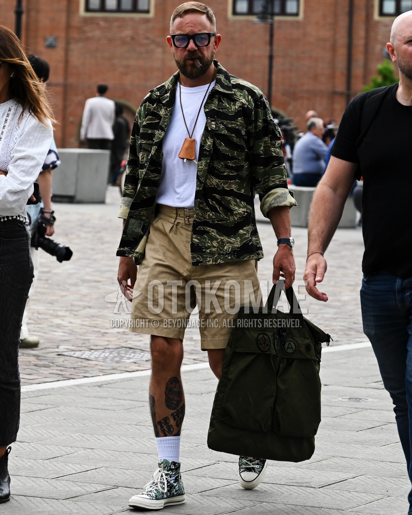 Men's spring summer autumn outfit with green camouflage military jacket, white plain t-shirt, beige plain short pants, white plain socks, green high-cut sneakers, black plain tote bag.