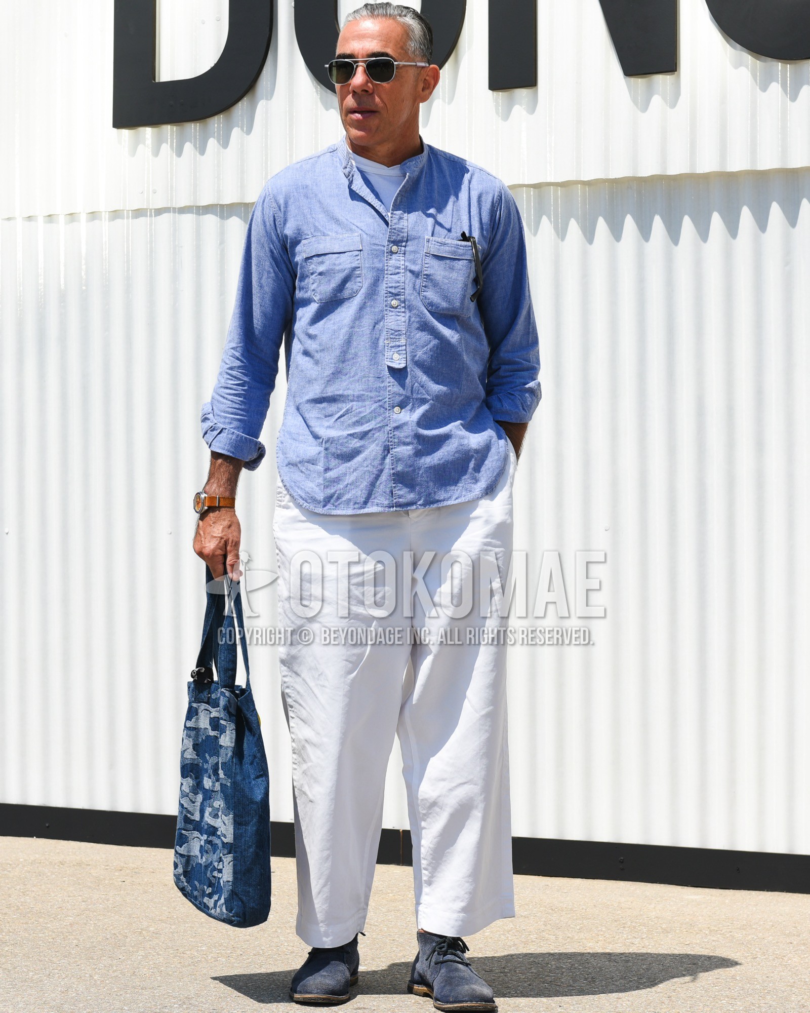 Men's spring summer outfit with silver plain sunglasses, white plain t-shirt, blue plain shirt, white plain cotton pants, navy chukka boots, navy bag tote bag.