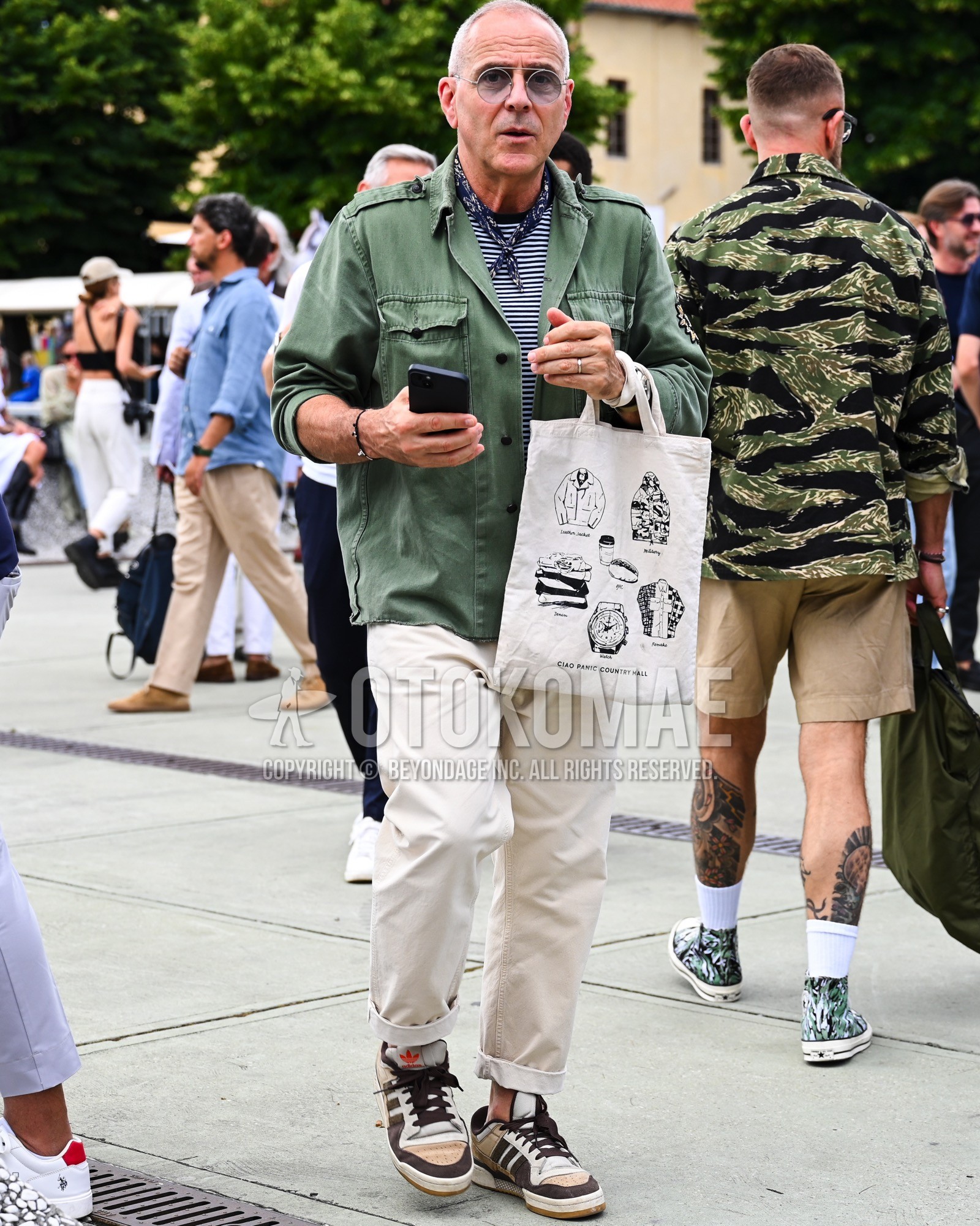 Men's spring summer autumn outfit with gray plain sunglasses, navy whole pattern bandana/neckerchief, olive green plain military jacket, navy horizontal stripes t-shirt, white plain chinos, beige low-cut sneakers, white bag tote bag.