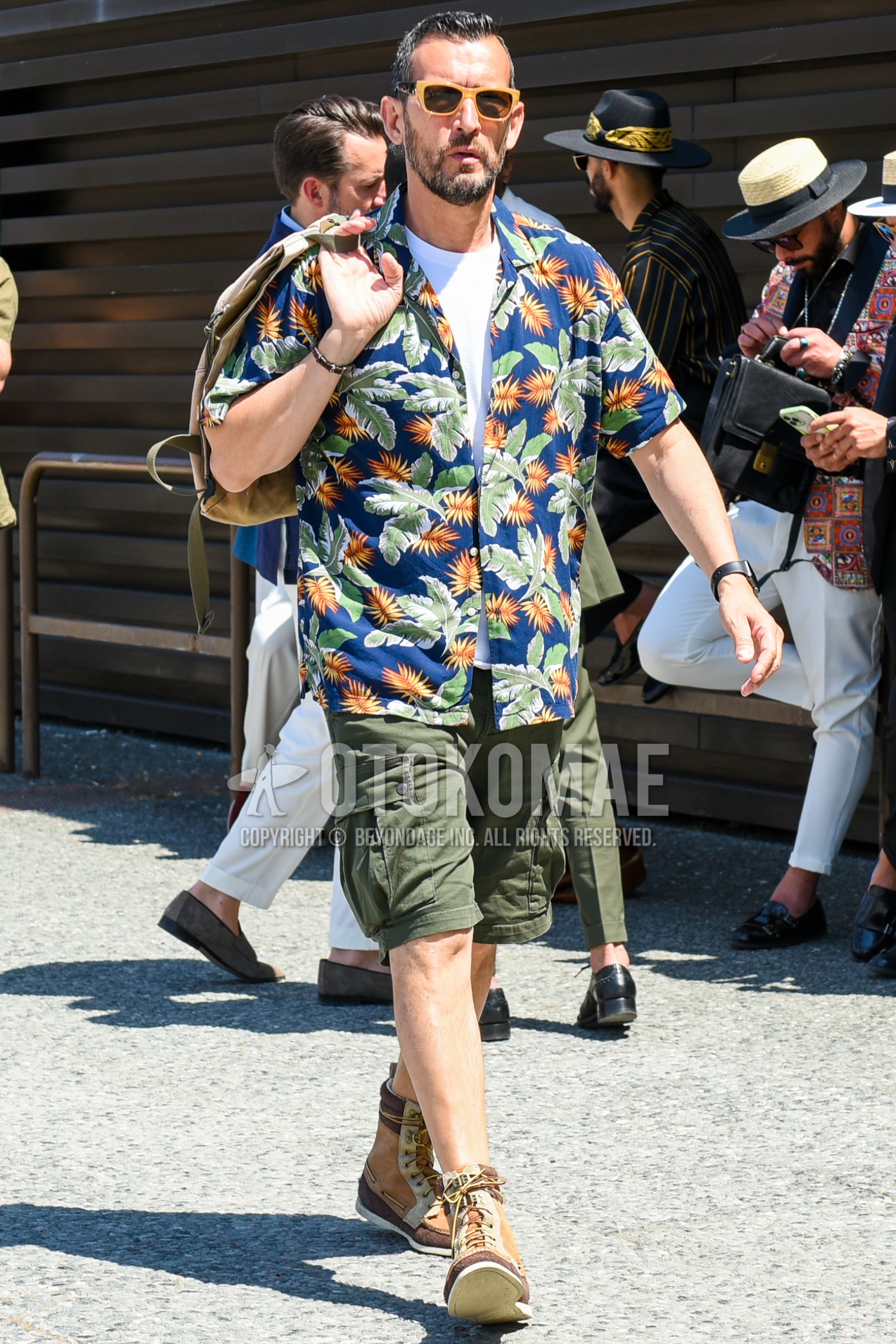Men's spring summer outfit with brown plain sunglasses, blue botanical shirt, olive green plain short pants, olive green plain cargo pants, brown  boots, beige plain briefcase/handbag.