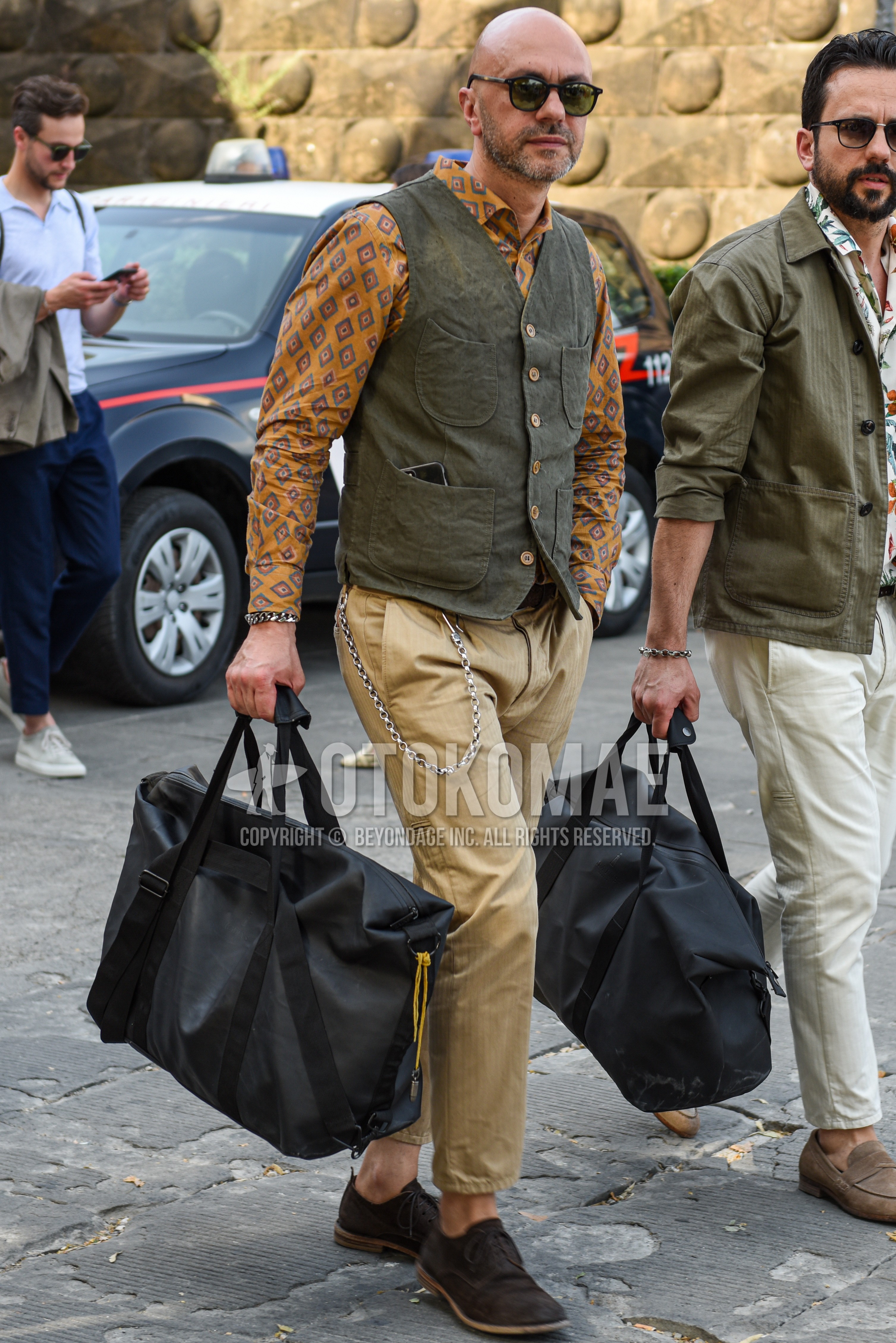 Men's spring summer outfit with black plain sunglasses, olive green plain gilet, orange tops/innerwear shirt, beige plain chinos, brown wing-tip shoes leather shoes.