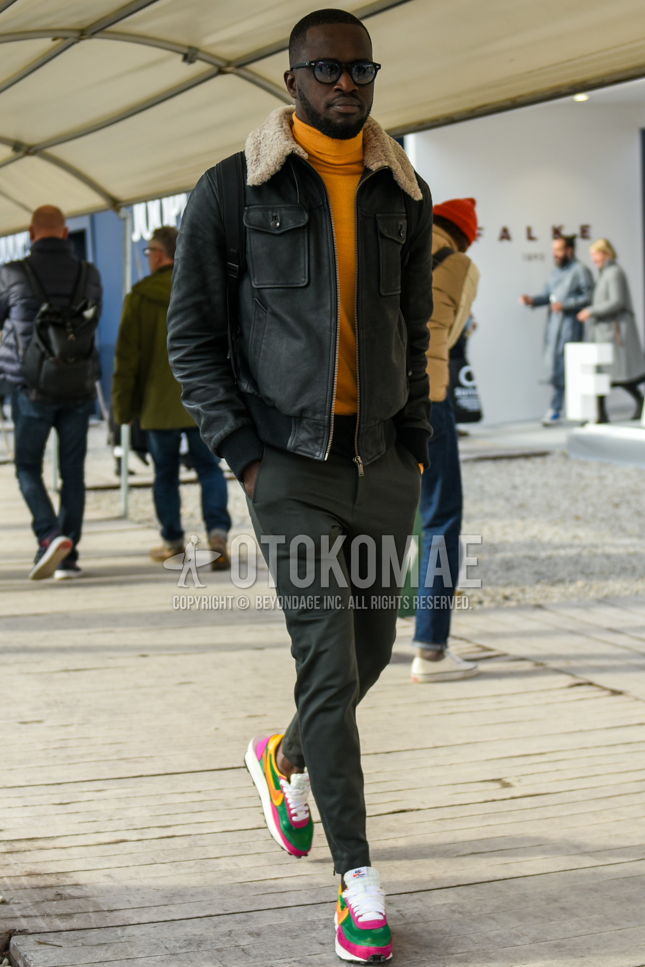 Men's autumn winter outfit with black plain sunglasses, dark gray plain leather jacket, dark gray plain military jacket, yellow plain turtleneck knit, gray plain slacks, multi-color low-cut sneakers.