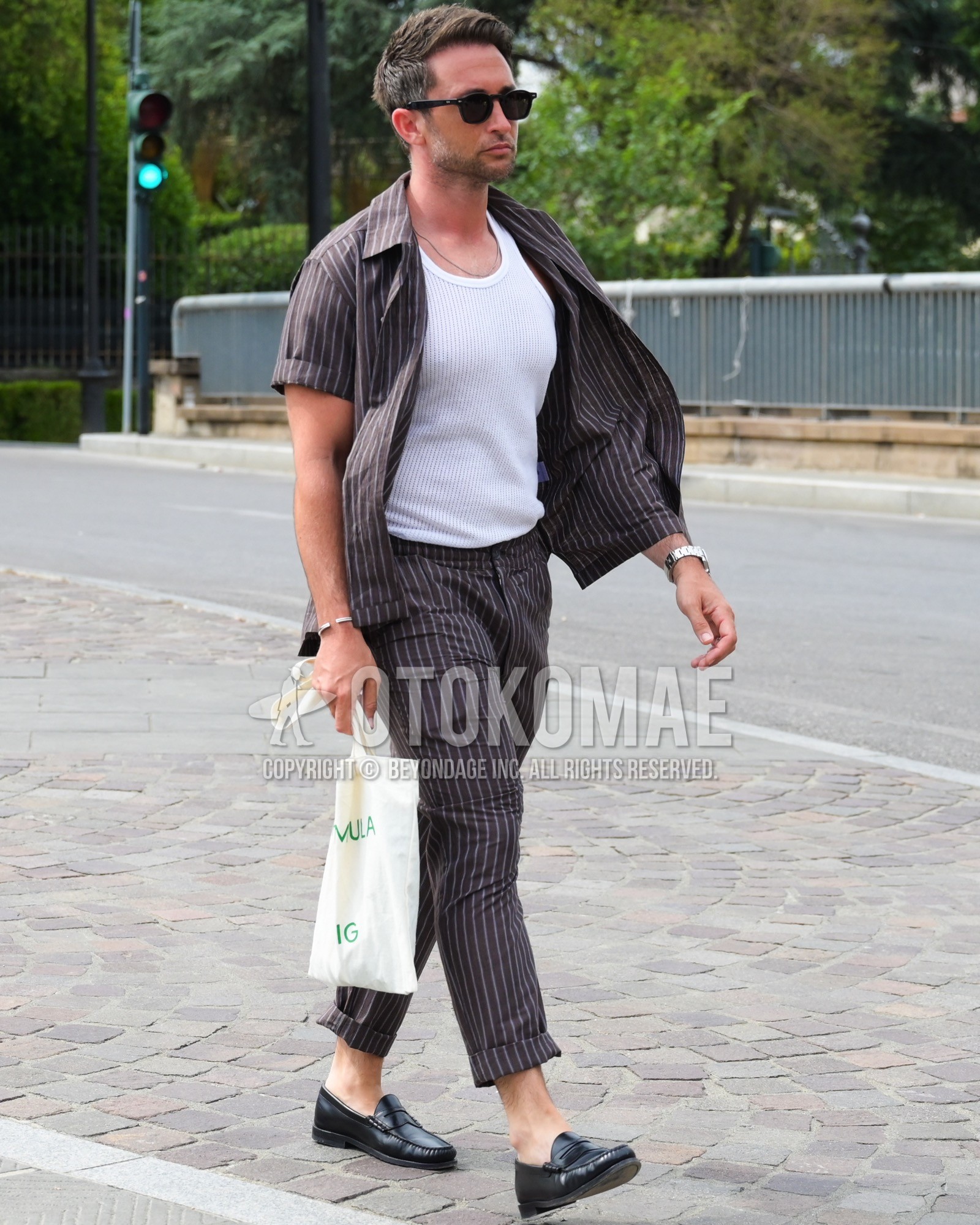 Men's spring summer outfit with black plain sunglasses, white plain tank top, black coin loafers leather shoes, white lettered tote bag, brown stripes suit.