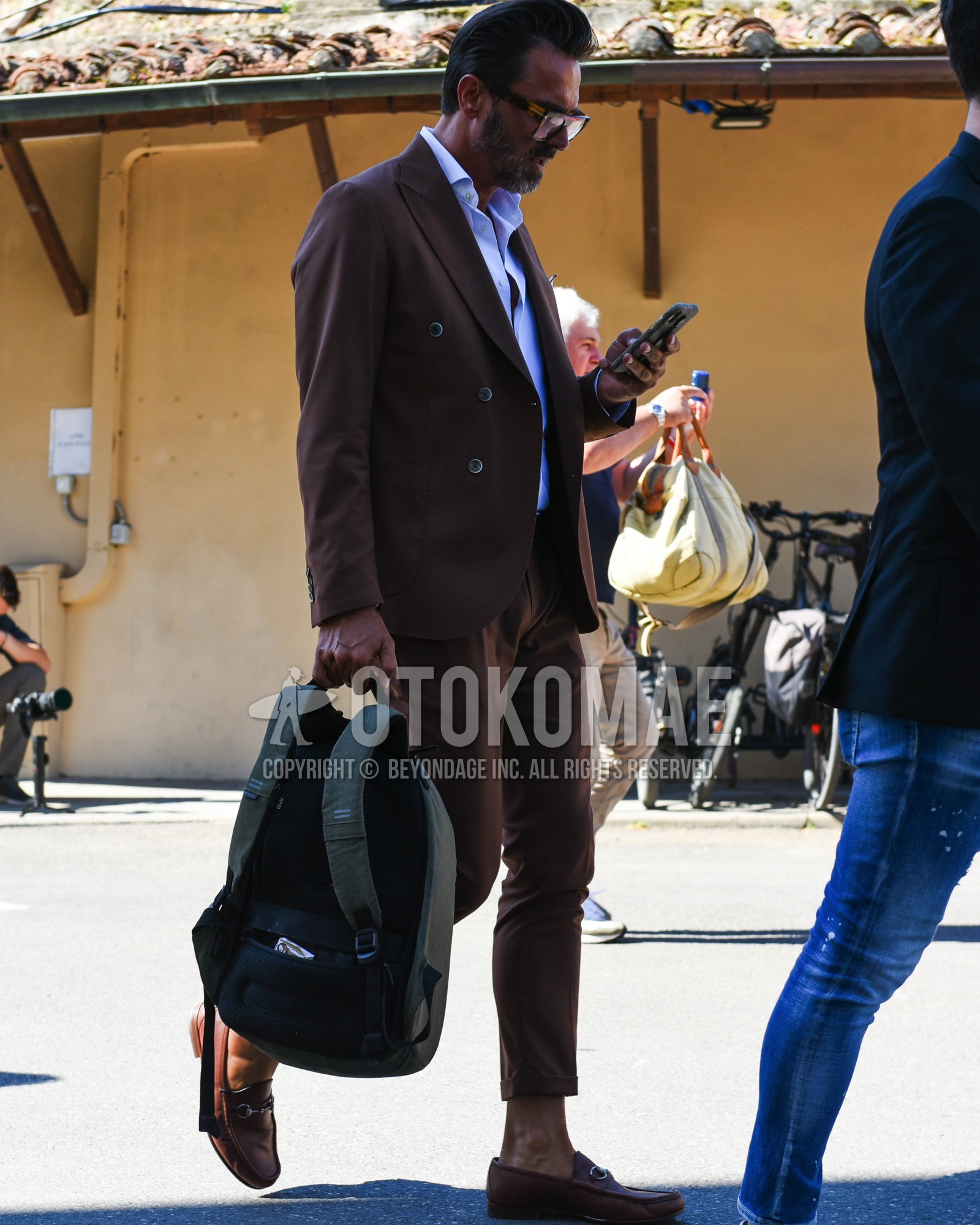 Men's spring summer outfit with yellow tortoiseshell sunglasses, white plain shirt, brown bit loafers leather shoes, orange plain backpack, brown plain suit.