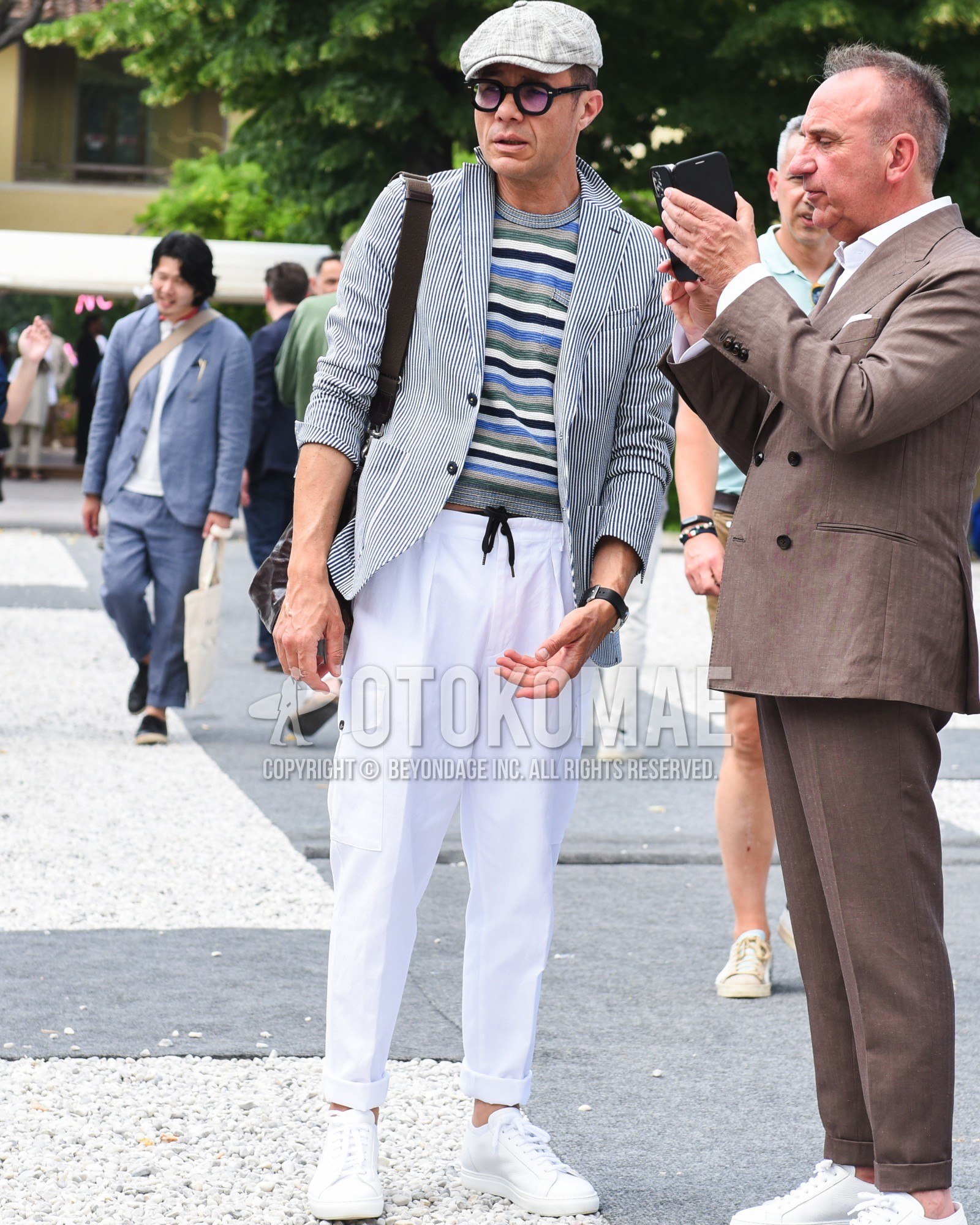 Men's spring summer outfit with gray check hunting cap, black plain glasses, navy white stripes tailored jacket, multi-color horizontal stripes t-shirt, white plain easy pants, white low-cut sneakers, brown plain shoulder bag.