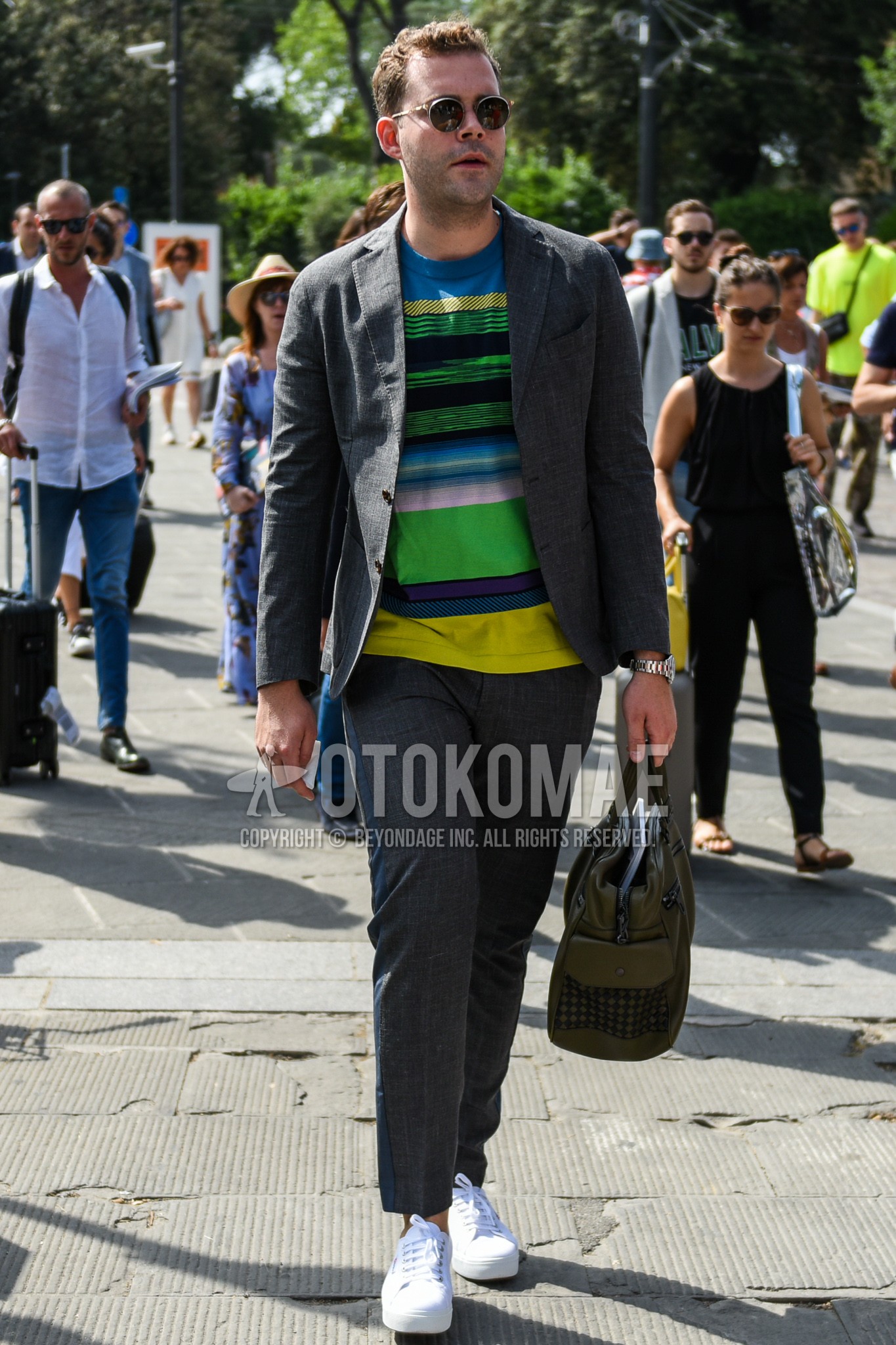 Men's spring summer autumn outfit with clear plain sunglasses, light blue yellow green horizontal stripes t-shirt, white low-cut sneakers, olive green plain briefcase/handbag.