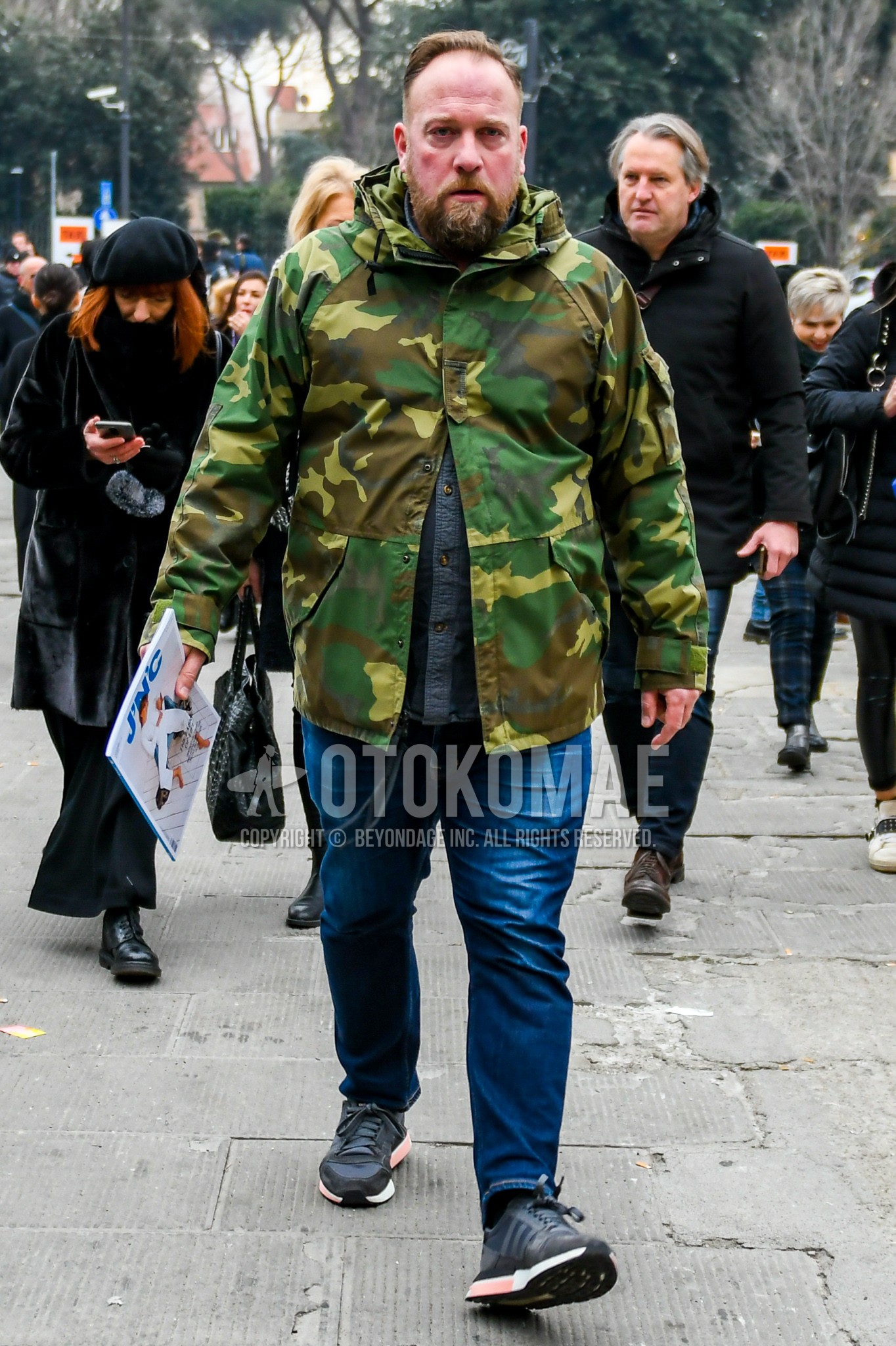 Men's autumn winter outfit with olive green camouflage hooded coat, gray plain shirt, blue plain denim/jeans, black low-cut sneakers.