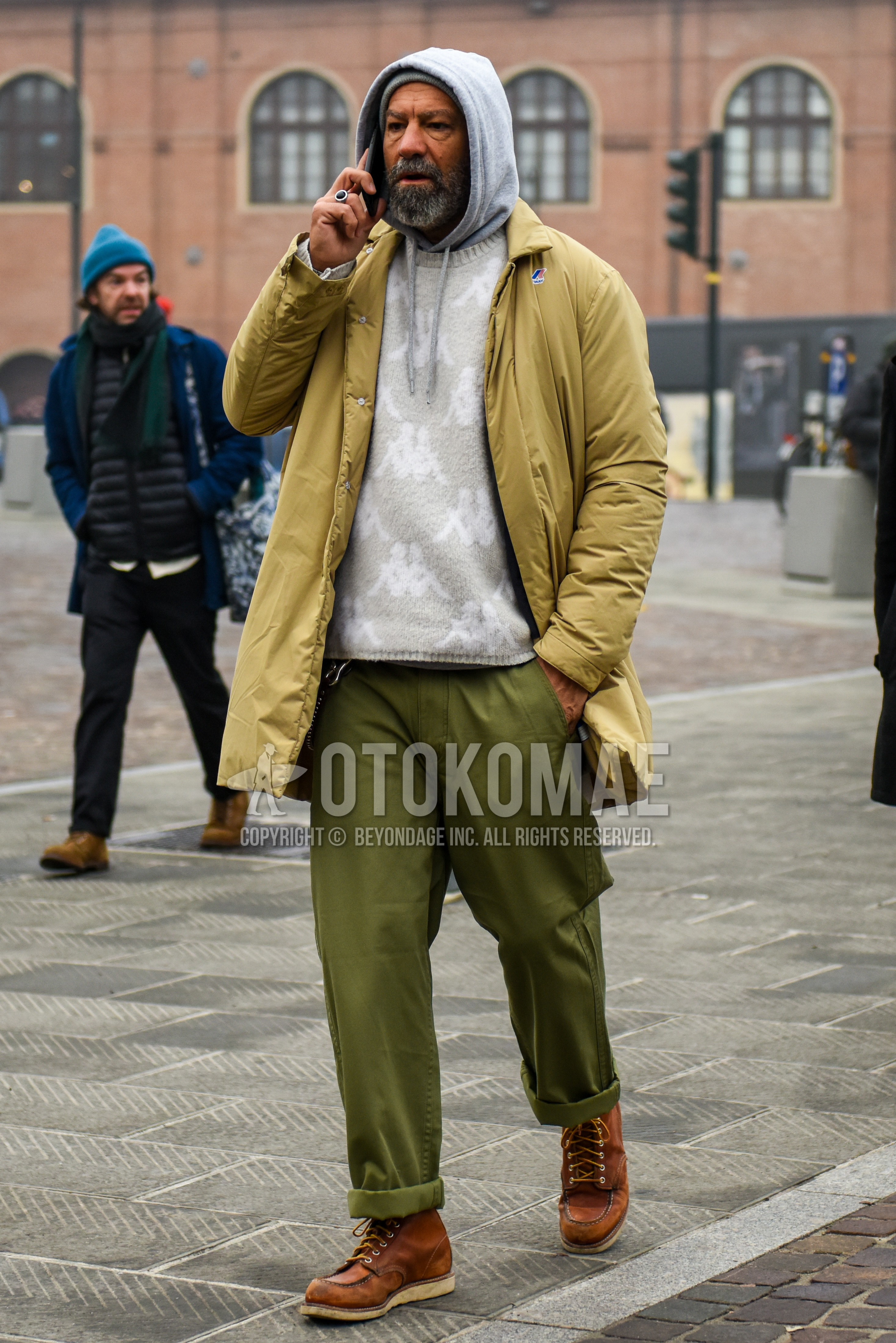 Men's winter outfit with beige plain stenkarrer coat, beige plain down jacket, gray graphic sweater, gray plain hoodie, olive green plain chinos, brown work boots.