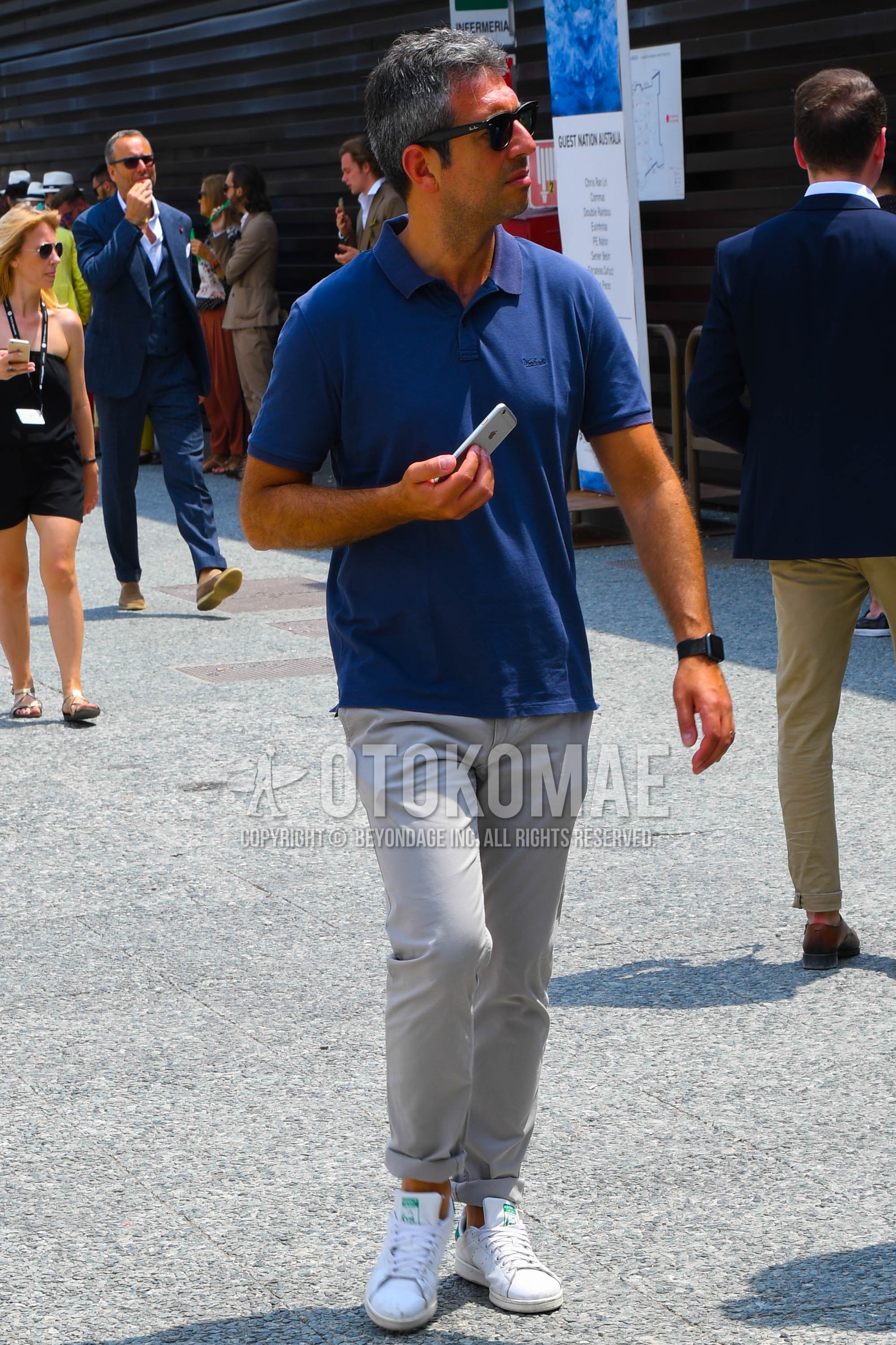 Men's summer outfit with plain sunglasses, blue plain polo shirt, beige plain chinos, white low-cut sneakers.