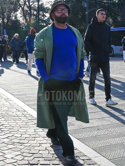 オリーブグリーンワンポイントのバケットハット、黒無地のメガネ、グリーン無地のチェスターコート、ブルー無地のセーター、黒無地のスラックス、黒プレーントゥの革靴を合わせた秋冬のメンズコーデ・着こなし。