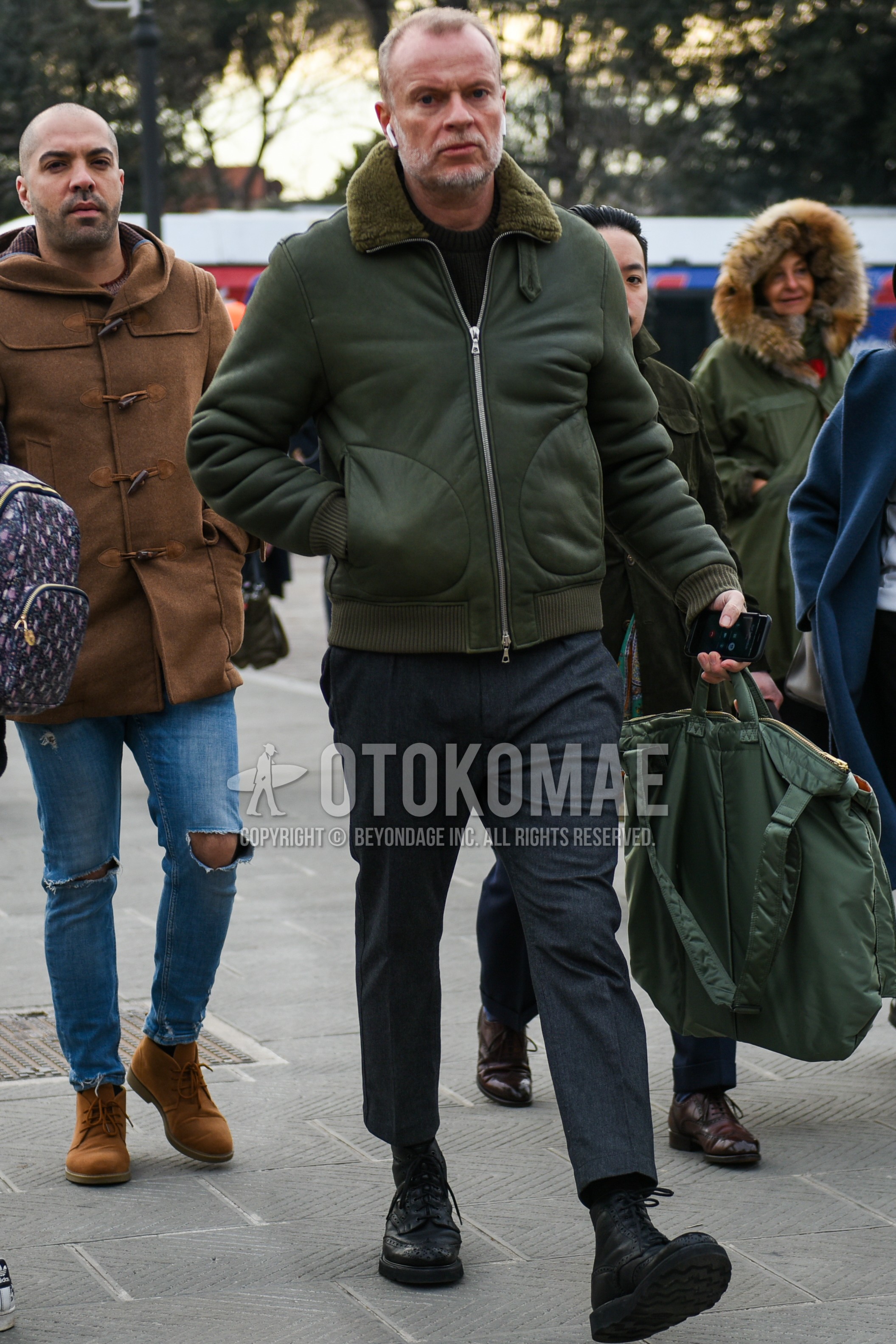 Men's autumn winter outfit with olive green plain military jacket, black plain sweatshirt, gray plain slacks, gray plain cropped pants, black plain socks, black  boots, green plain briefcase/handbag.