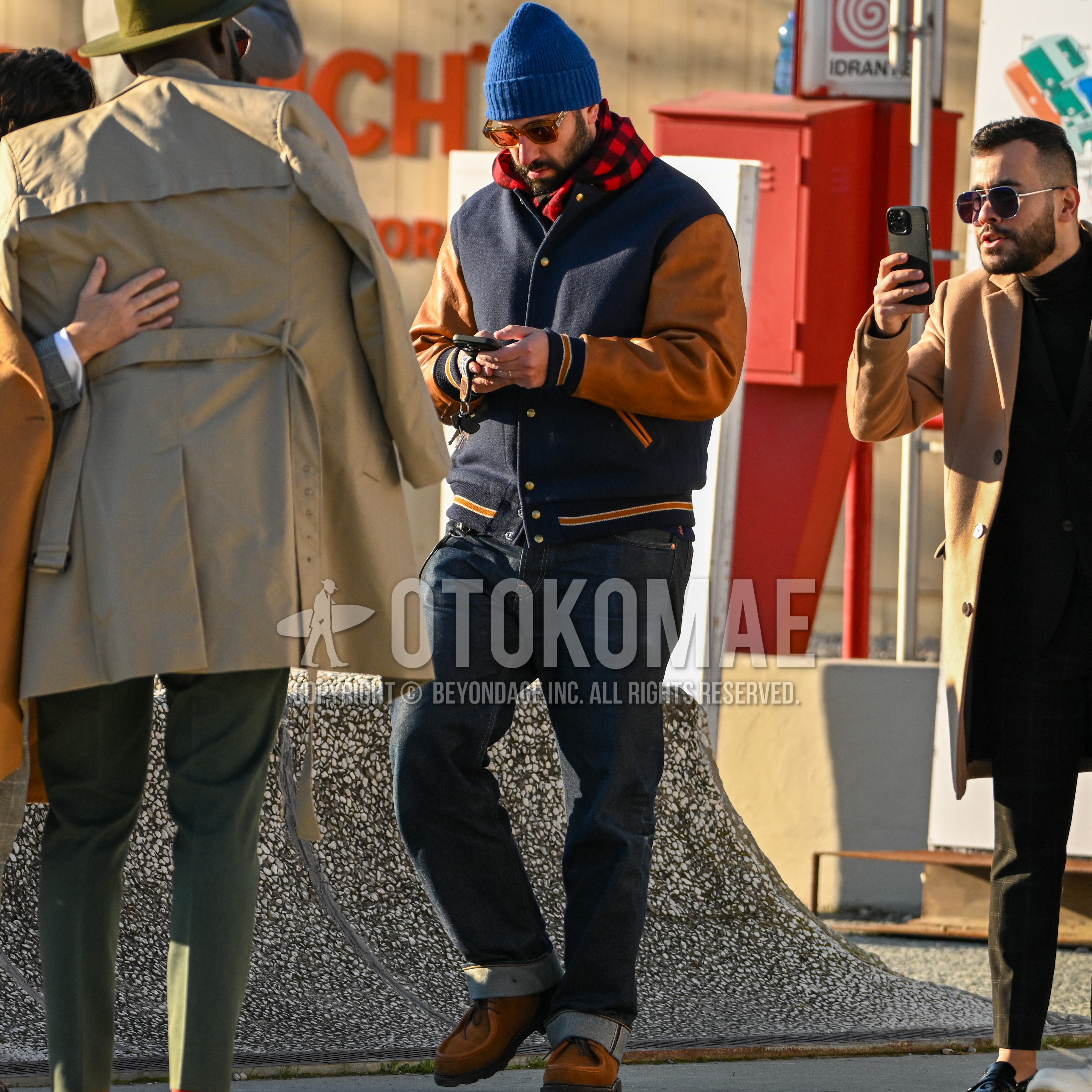 Men's autumn winter outfit with blue plain knit cap, clear plain sunglasses, red black check scarf, navy brown outerwear stadium jacket, plain denim/jeans, brown  loafers leather shoes.