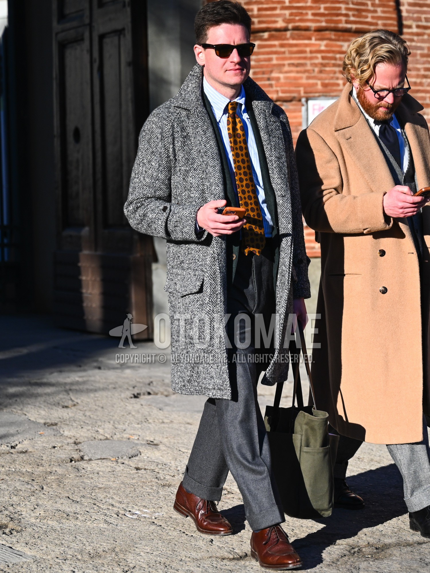 Men's autumn winter outfit with brown tortoiseshell sunglasses, gray herringbone ulster coat, green herringbone tailored jacket, white blue stripes shirt, gray plain slacks, brown u-tip shoes leather shoes, olive green plain tote bag, yellow small crest necktie.