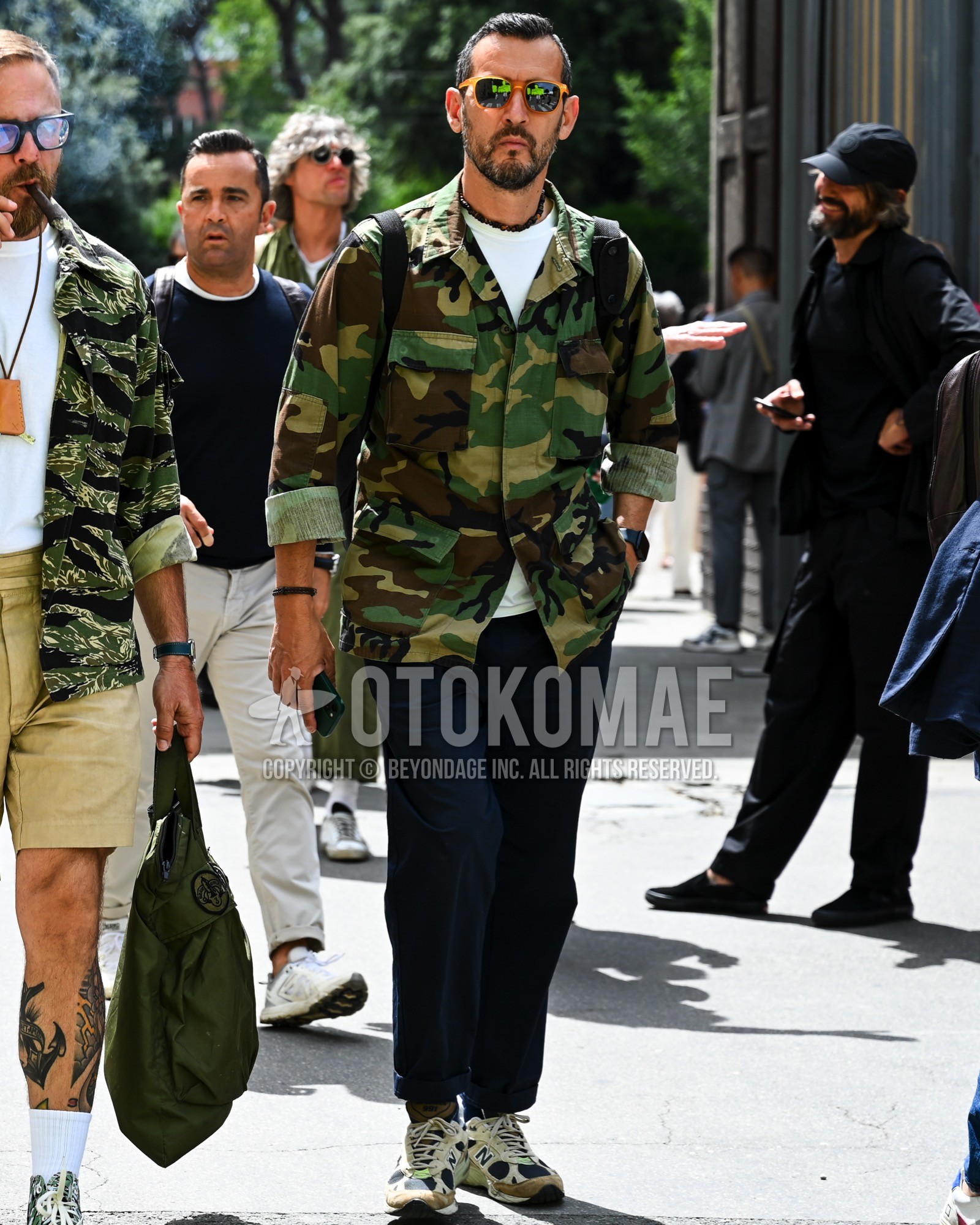 Men's spring summer autumn outfit with green plain sunglasses, olive green camouflage military jacket, white plain t-shirt, navy plain chinos, white low-cut sneakers.