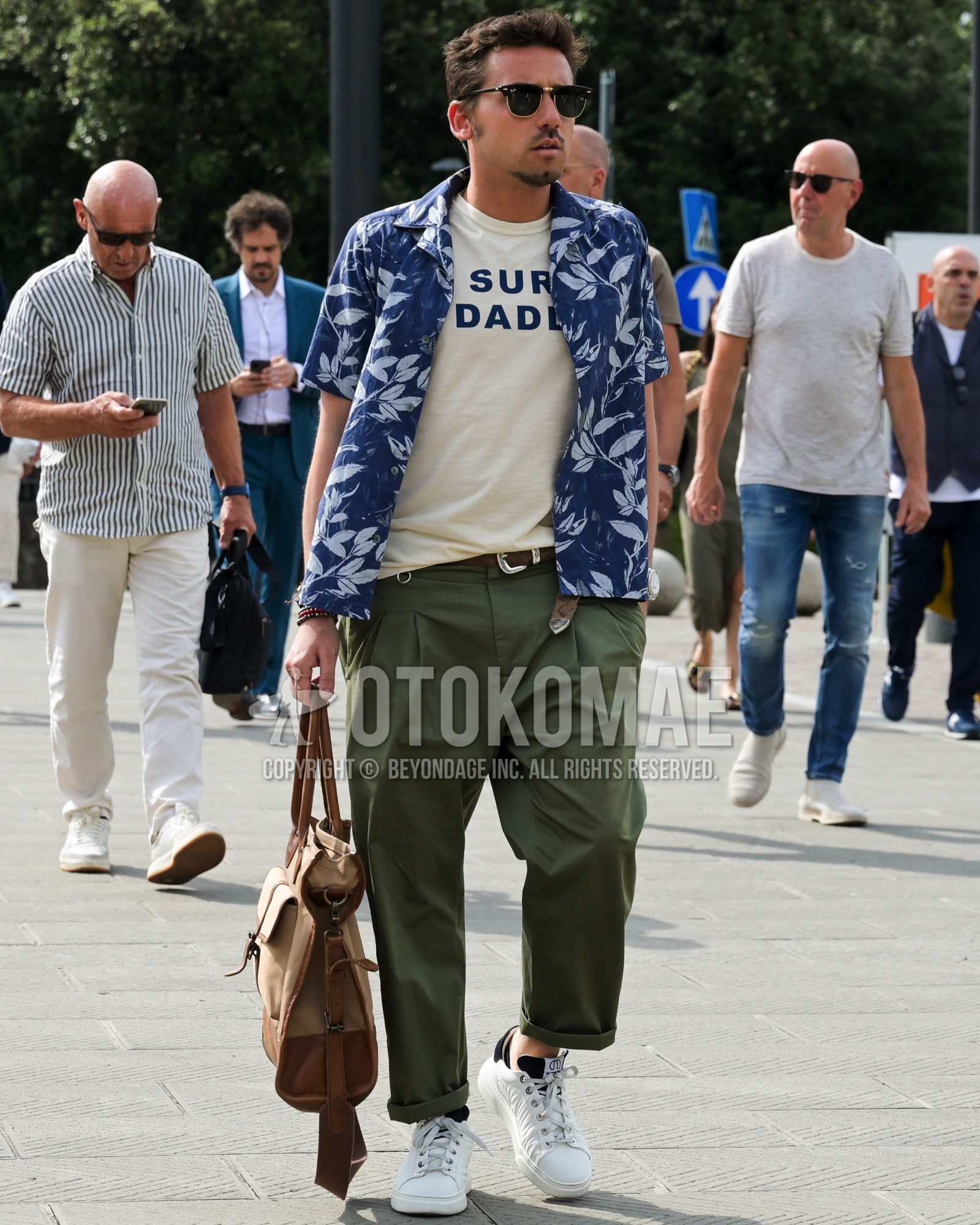 Men's spring summer outfit with black plain sunglasses, navy whole pattern shirt, beige lettered t-shirt, brown plain leather belt, olive green plain chinos, white low-cut sneakers, brown plain briefcase/handbag.