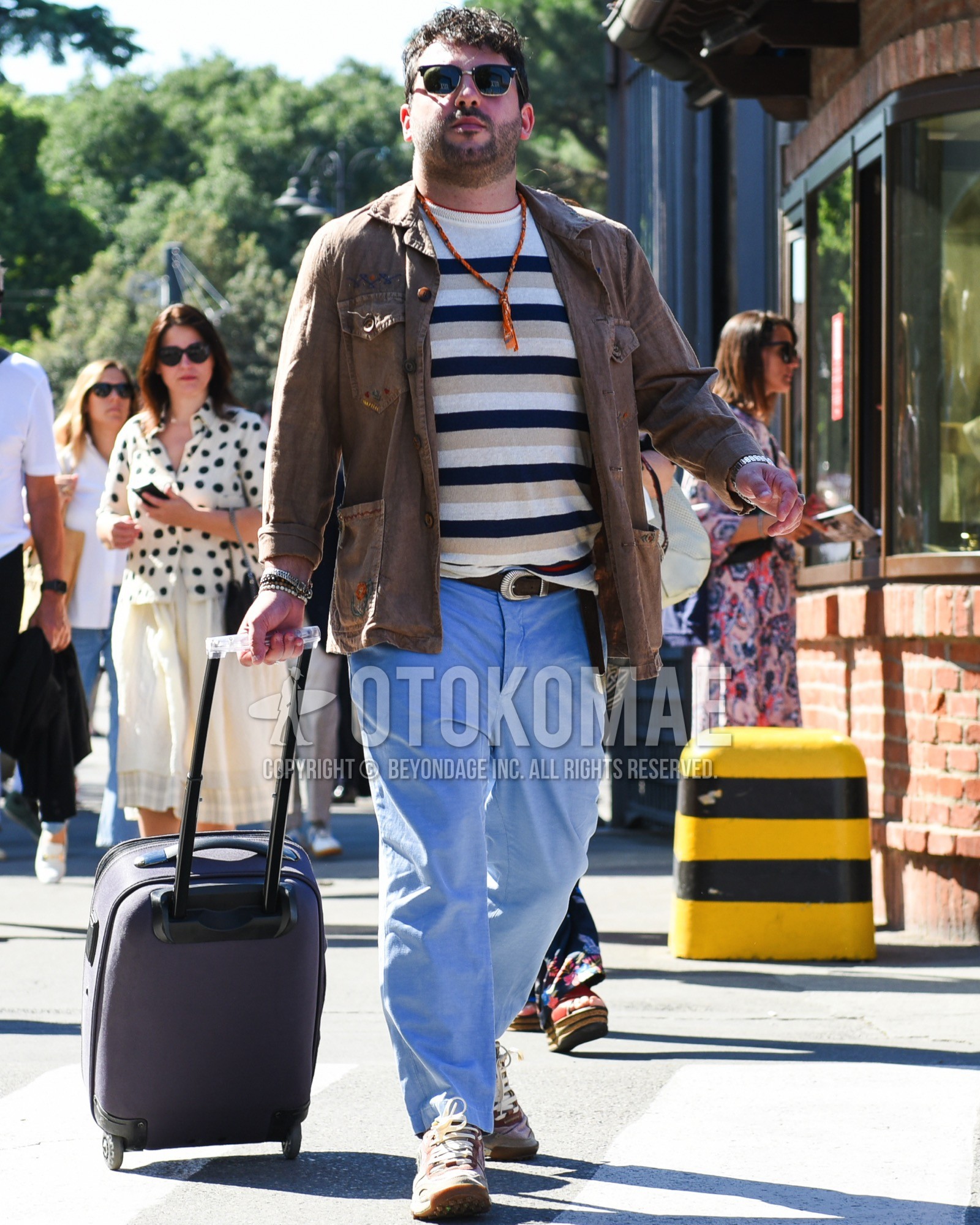 Men's spring summer outfit with black plain sunglasses, brown plain field jacket/hunting jacket, white beige navy horizontal stripes t-shirt, brown plain leather belt, blue plain chinos, brown low-cut sneakers, gray plain suitcase.