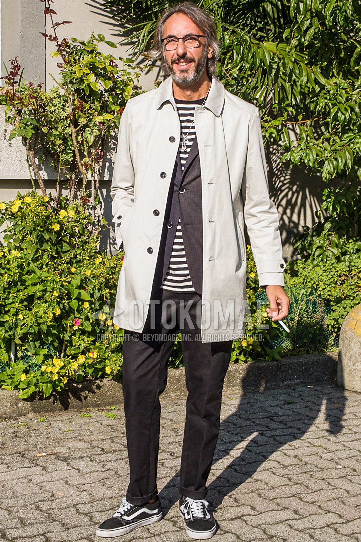 Men's spring autumn outfit with black plain glasses, beige plain stenkarrer coat, gray stripes tailored jacket, white black horizontal stripes t-shirt, gray plain slacks, black low-cut sneakers.